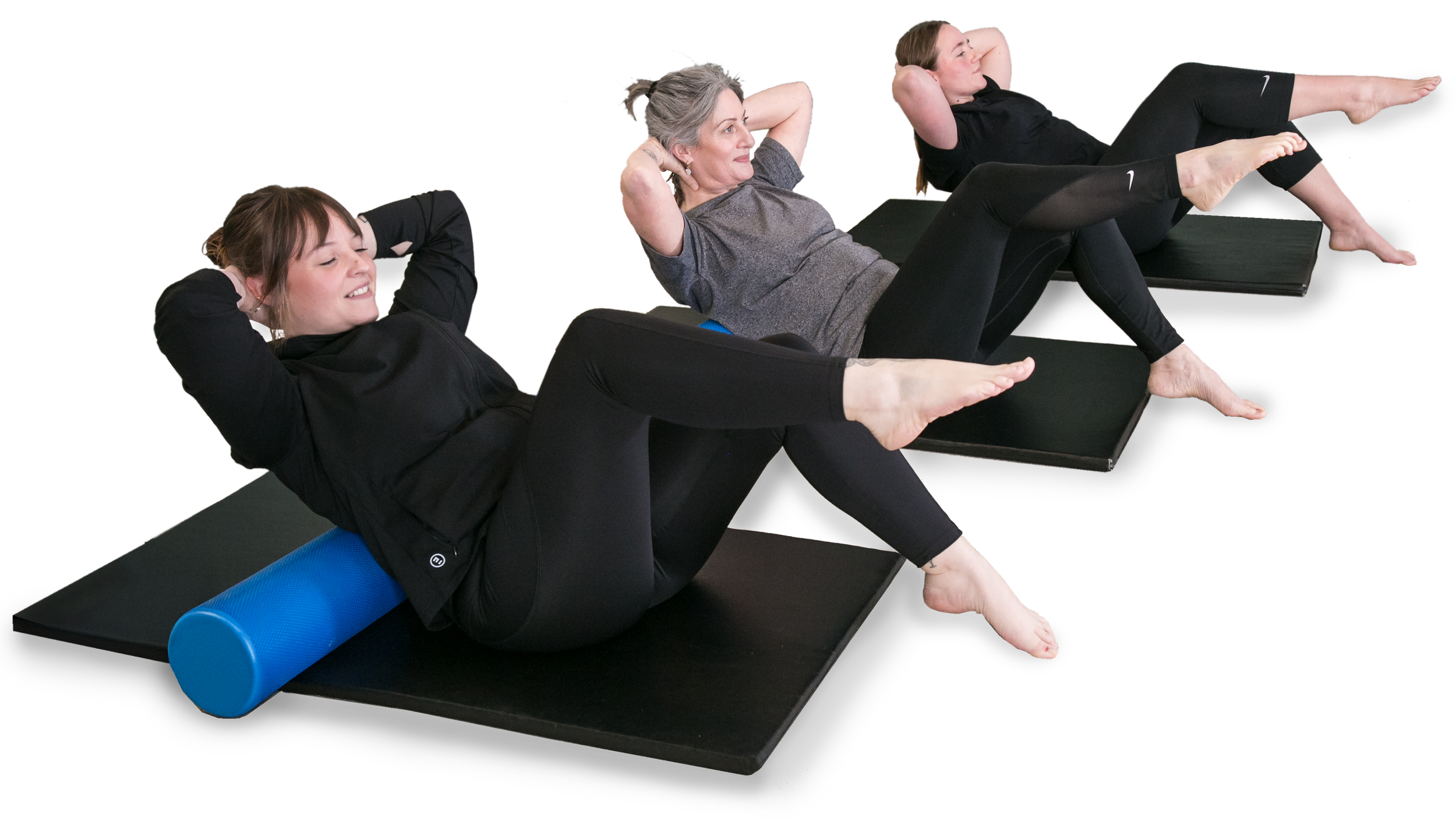 Three women exercise together on black mats, performing abdominal crunches with legs raised and hands behind their heads, using foam rollers under their backs, in a fitness class.