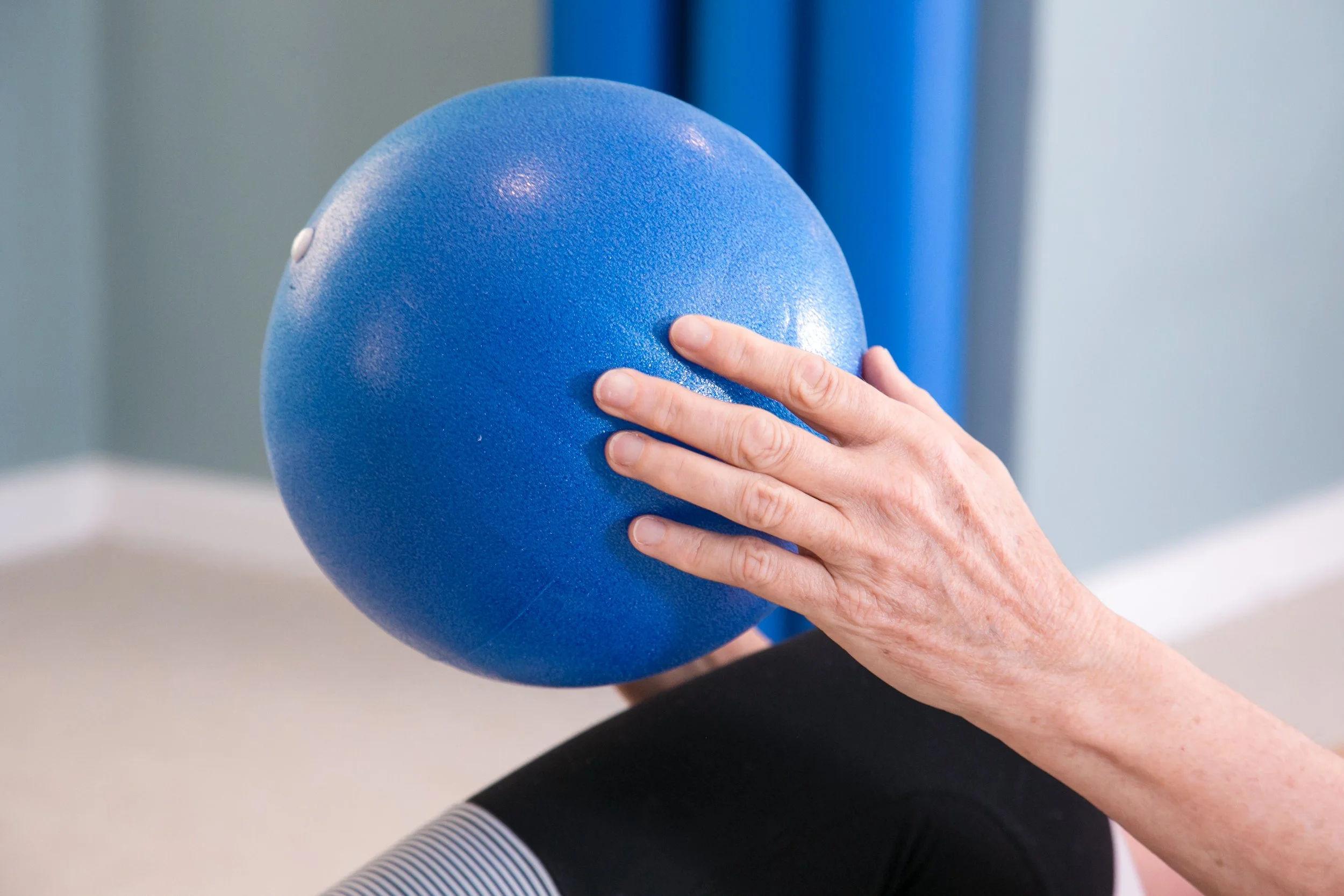 Close-up of a hand gripping a blue exercise ball, with a background of a light blue and darker blue curtain.