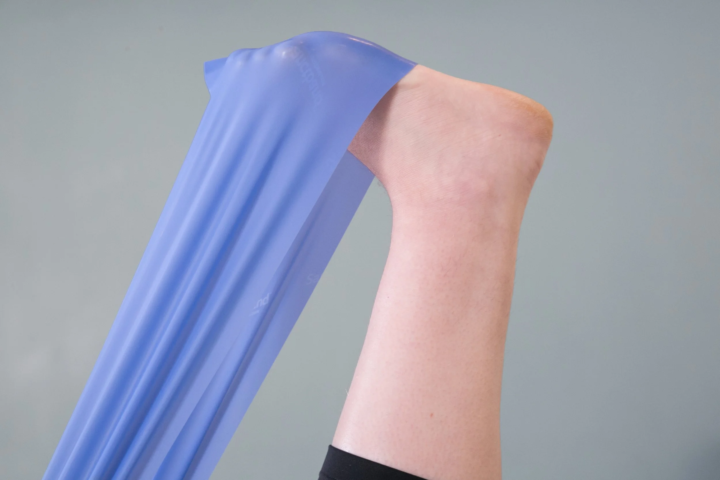 Close-up of a person with light skin, wearing a black wristband, holding a blue resistance band. The resistance band is stretched and held against a plain, light gray background.