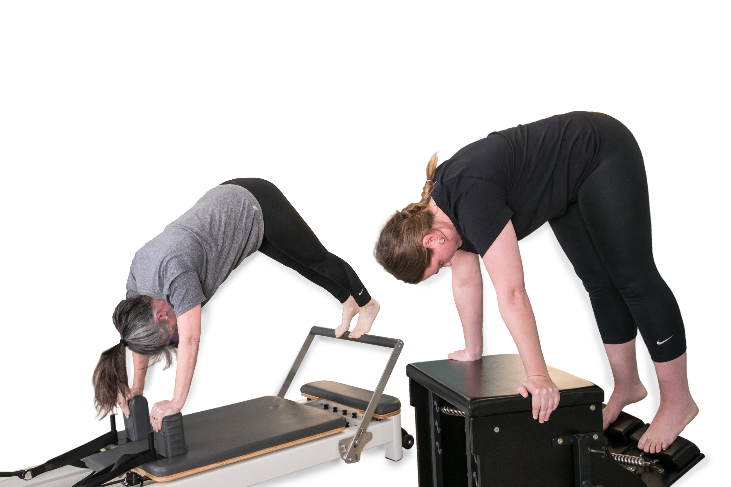 Two women practicing yoga in a downward dog pose, with one on a reformer machine and the other on a Pilates box, against a white background.