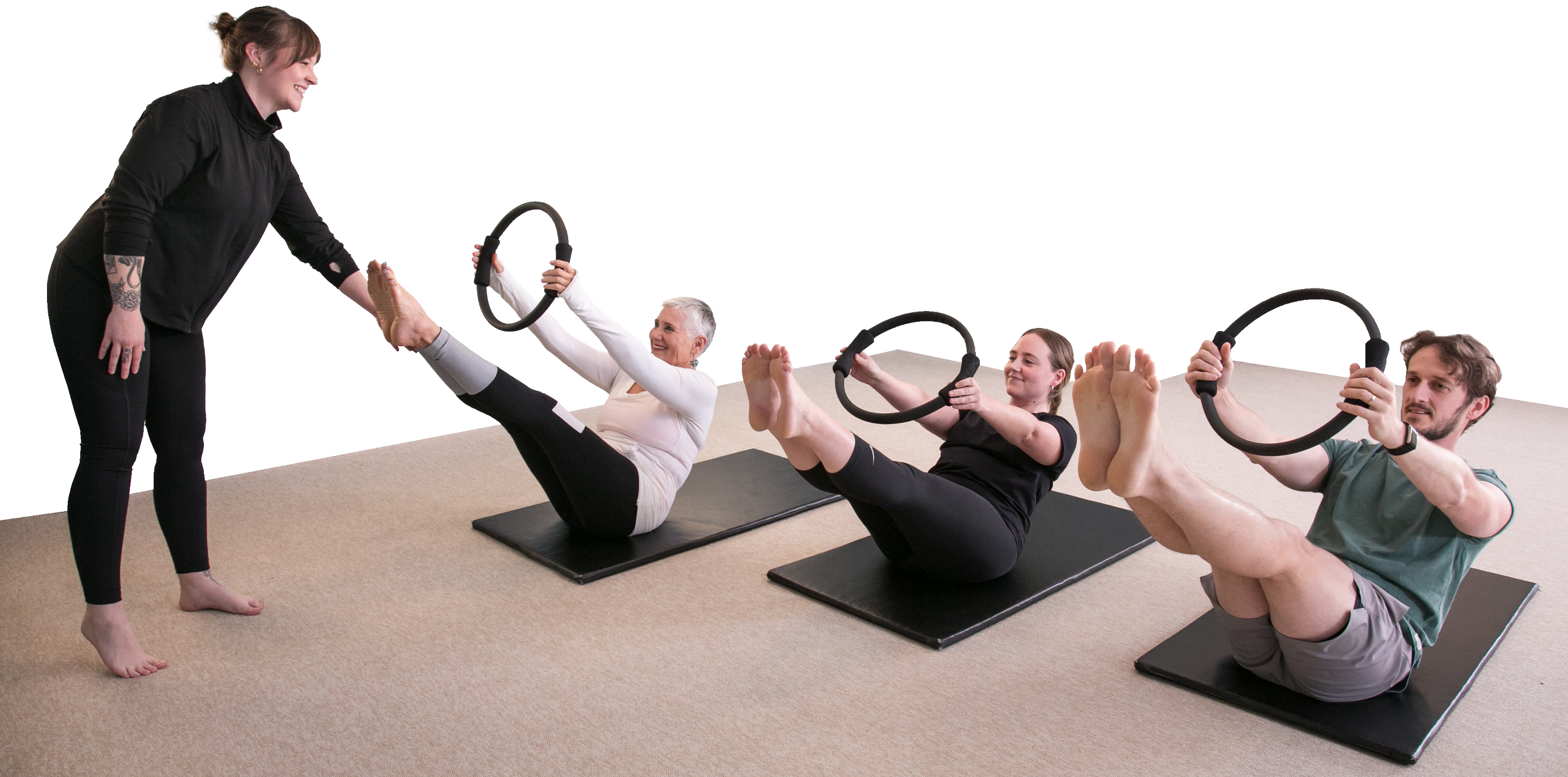 Instructor assisting three people lying on pilates mats, with raised legs and upper bodies, each holding a pilates ring out in front of them.