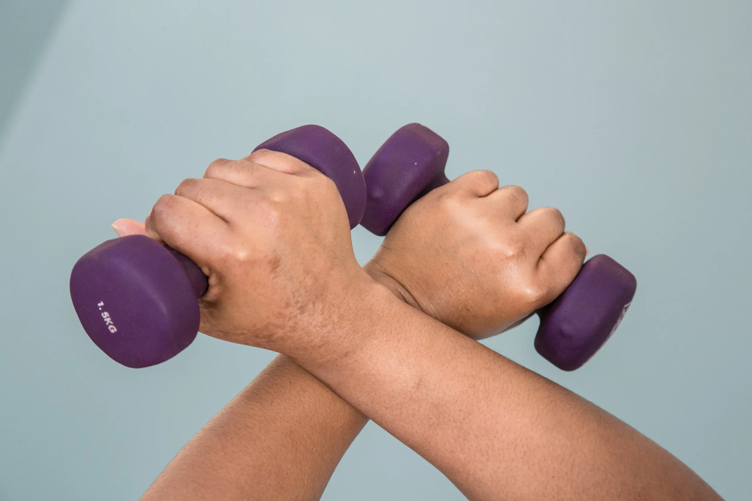 Two hands gripping purple dumbbells crossed over each other against a light blue background.