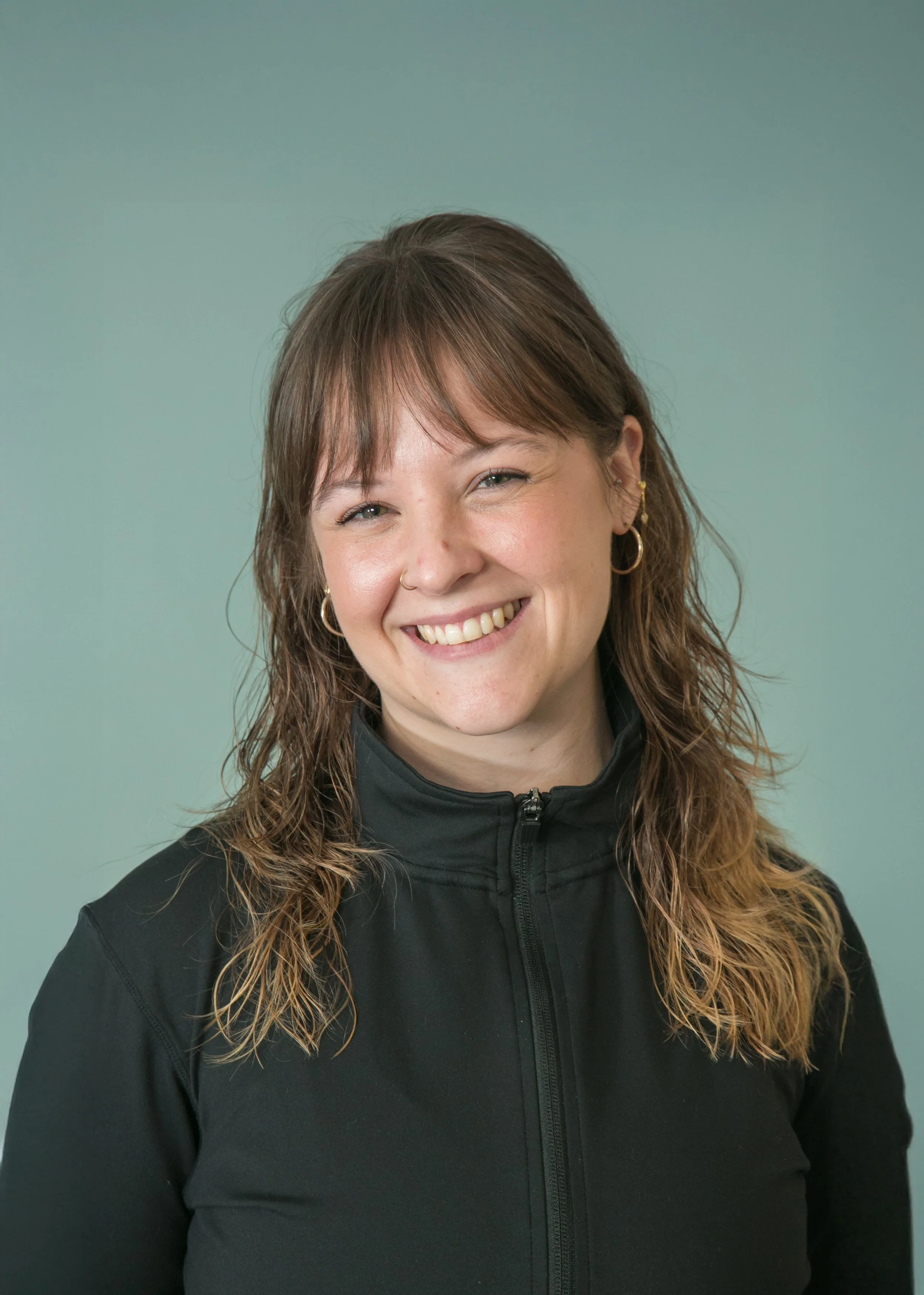 A smiling woman with light skin, long wavy brown hair, wearing hoop earrings and a nose ring, dressed in a black zip-up jacket, against a light green background.
