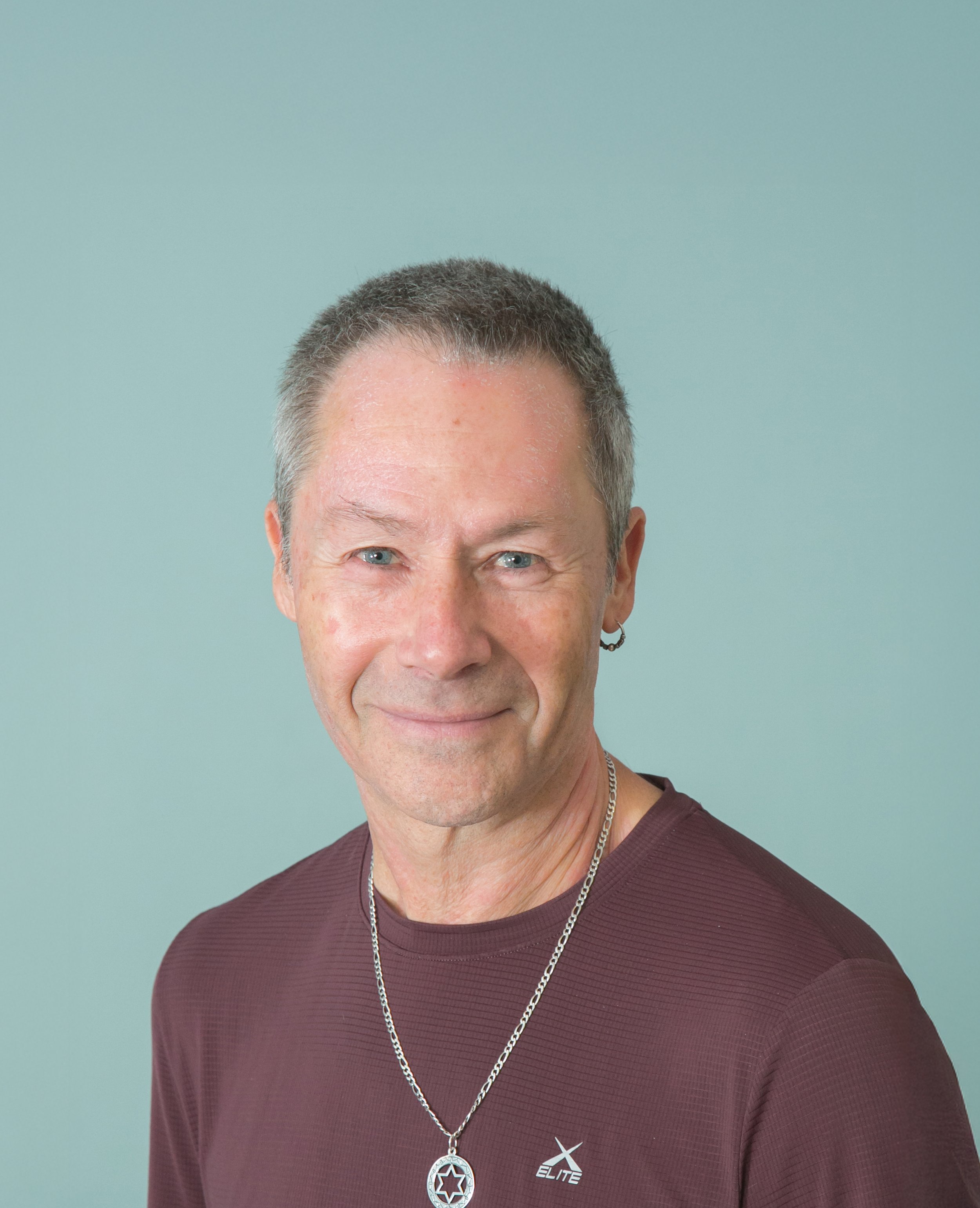 Half of a smiling man with short hair, blue eyes, and light skin, wearing a maroon shirt with a star-shaped pendant necklace and an 'ELITE' logo, against a plain light green background.