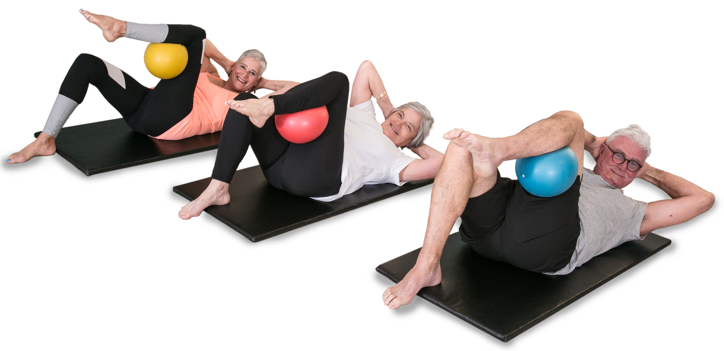 Three elderly individuals lying on exercise mats, performing abdominal exercises with small exercise balls placed under their legs. They are smiling and appear to be in a fitness class.