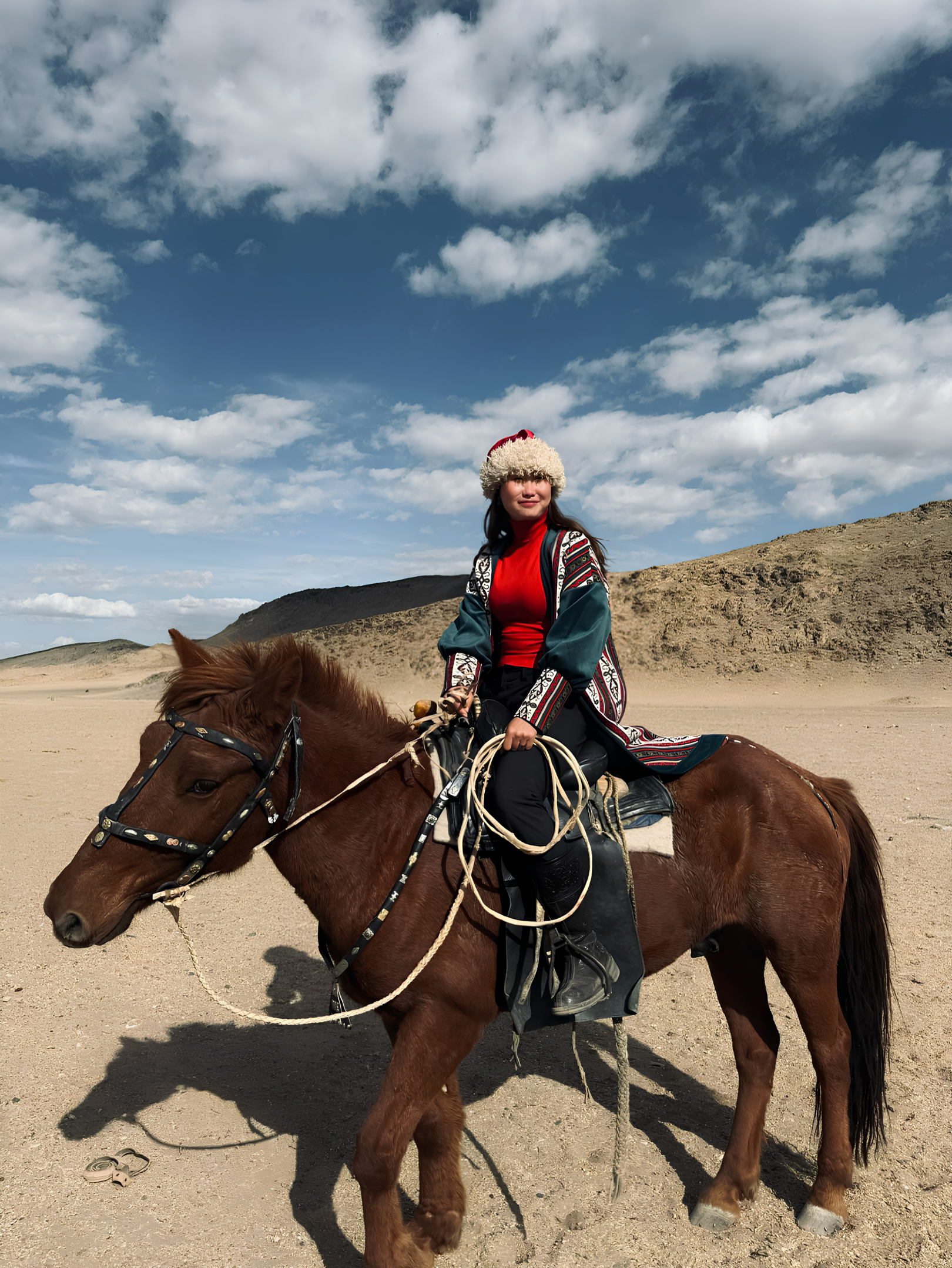 A woman wearing winter clothing, including a fluffy hat and a red shirt, is riding a brown horse in a desert landscape under a blue sky with scattered clouds.