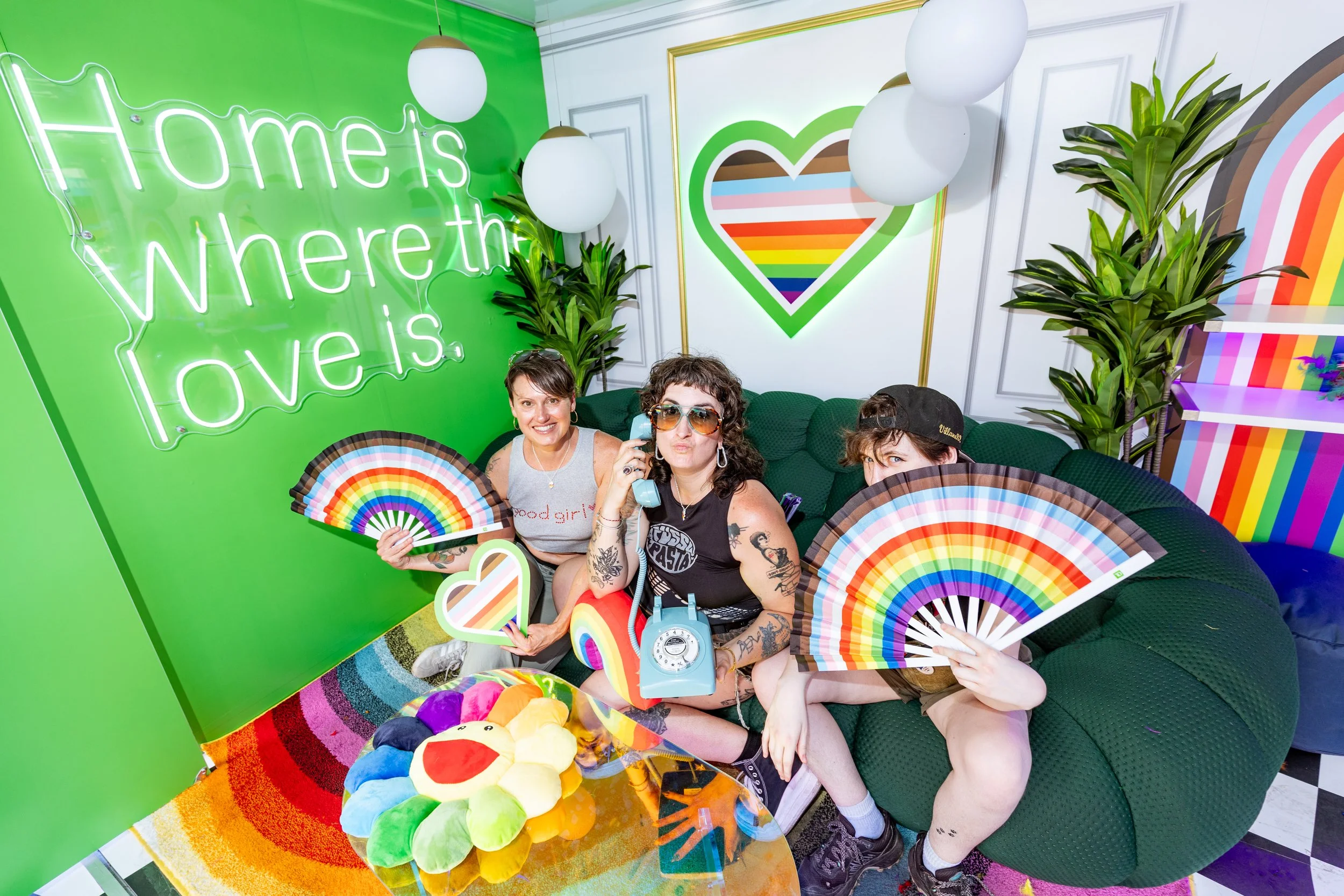 Group of three people sitting on a green couch in a colorful, rainbow-themed room with rainbow-colored fans, rainbow plush toys, and rainbow decor, including neon sign that says 'Home is where the love is' and rainbow art on the wall for TD Bank.
