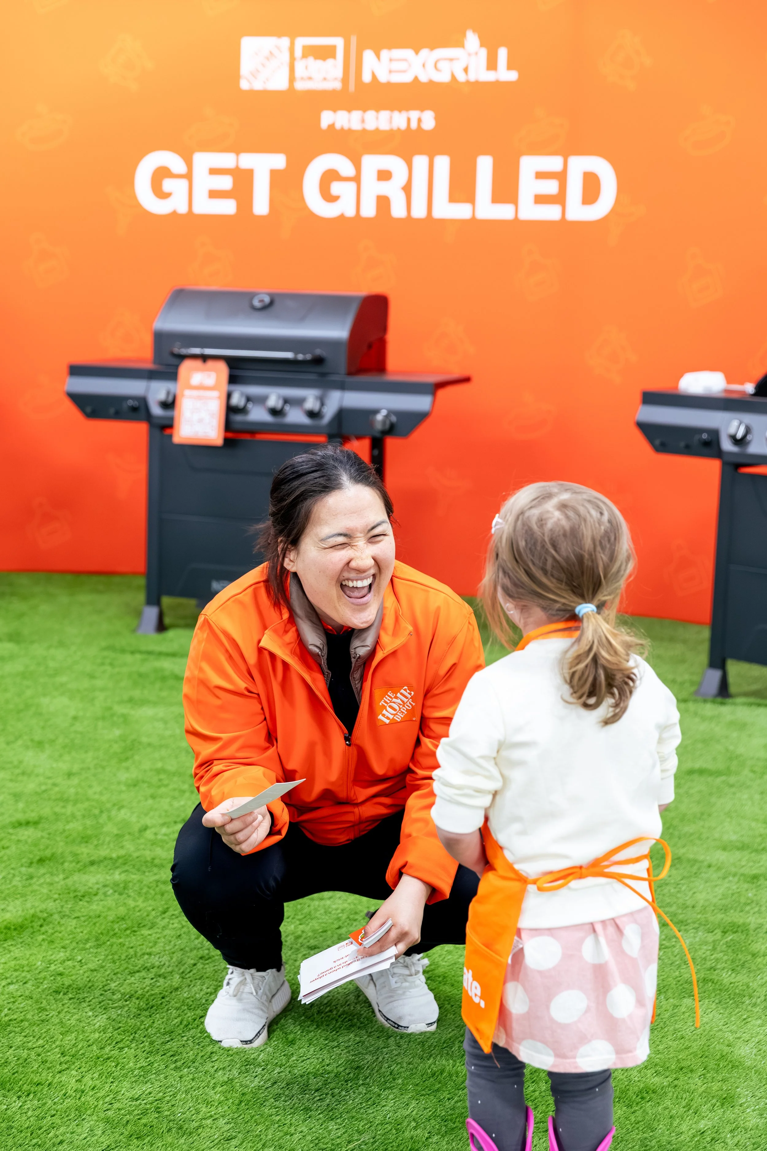 A woman kneeling and smiling, talking to a young girl who is holding some papers. The woman is wearing an orange jacket, and the girl is dressed in a white jacket and pink polka dot skirt with pink rain boots. They are at a Home Depot event.