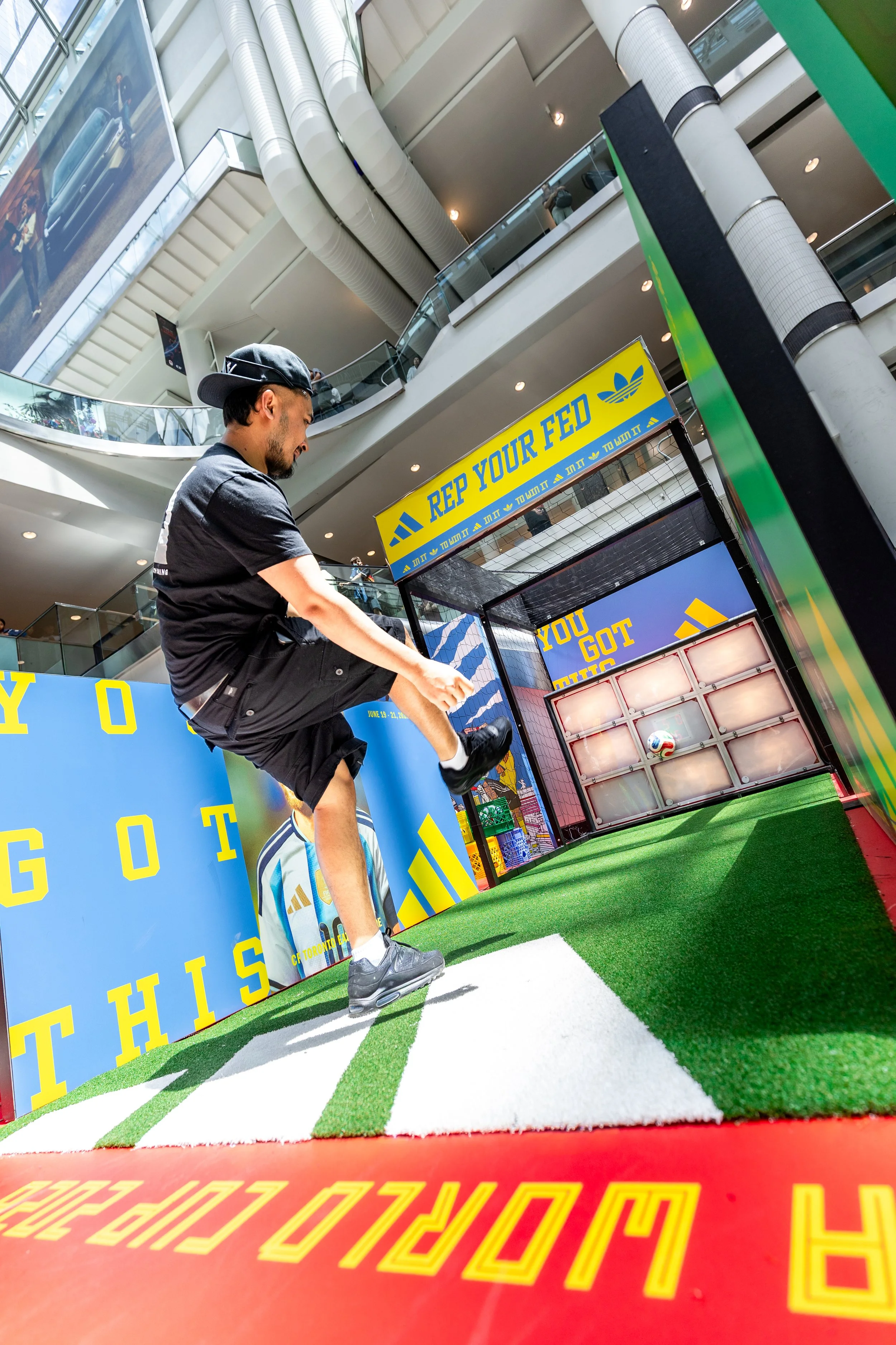 A person wearing a black cap and black t-shirt playing a soccer game at an indoor sports area, with banners and advertisements in the background promoting Adidas.