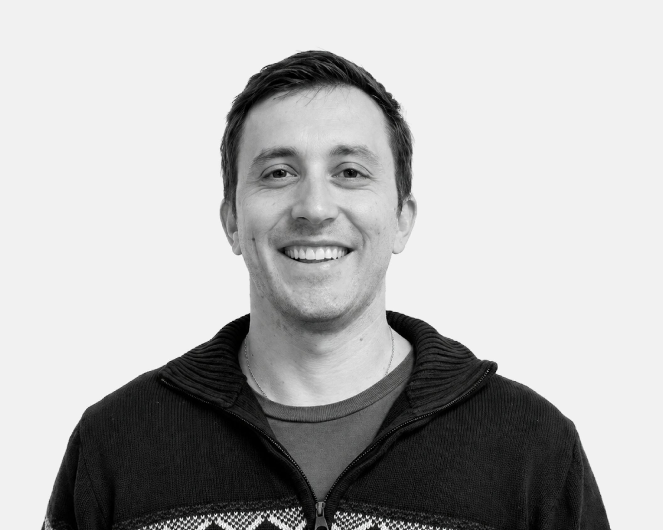 Black and white photo of a person with short hair, smiling, wearing a zip-up hoodie with a patterned shirt underneath.