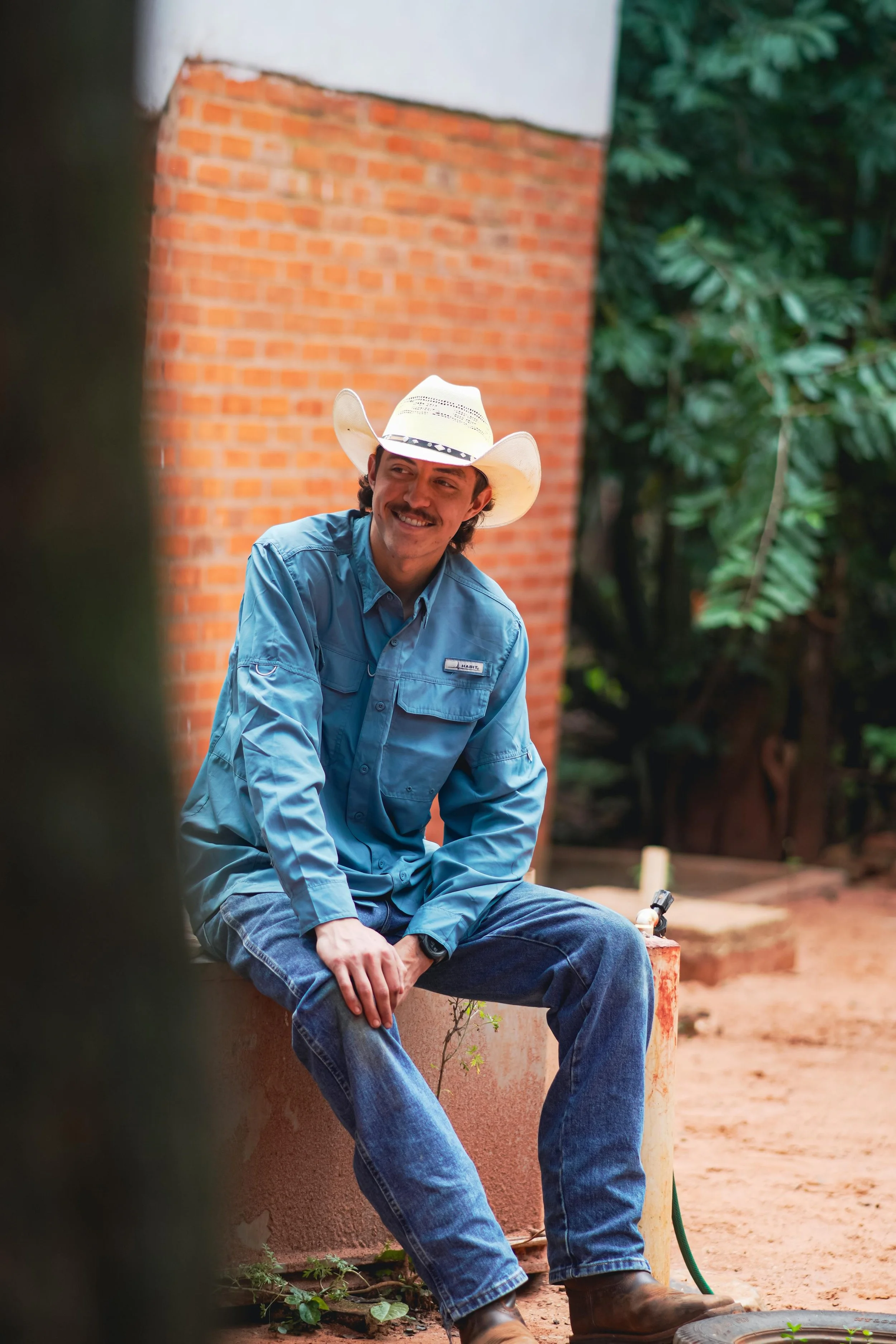 A man wearing a cowboy hat and blue shirt sits outdoors in front of a brick wall, smiling, with green foliage in the background.
