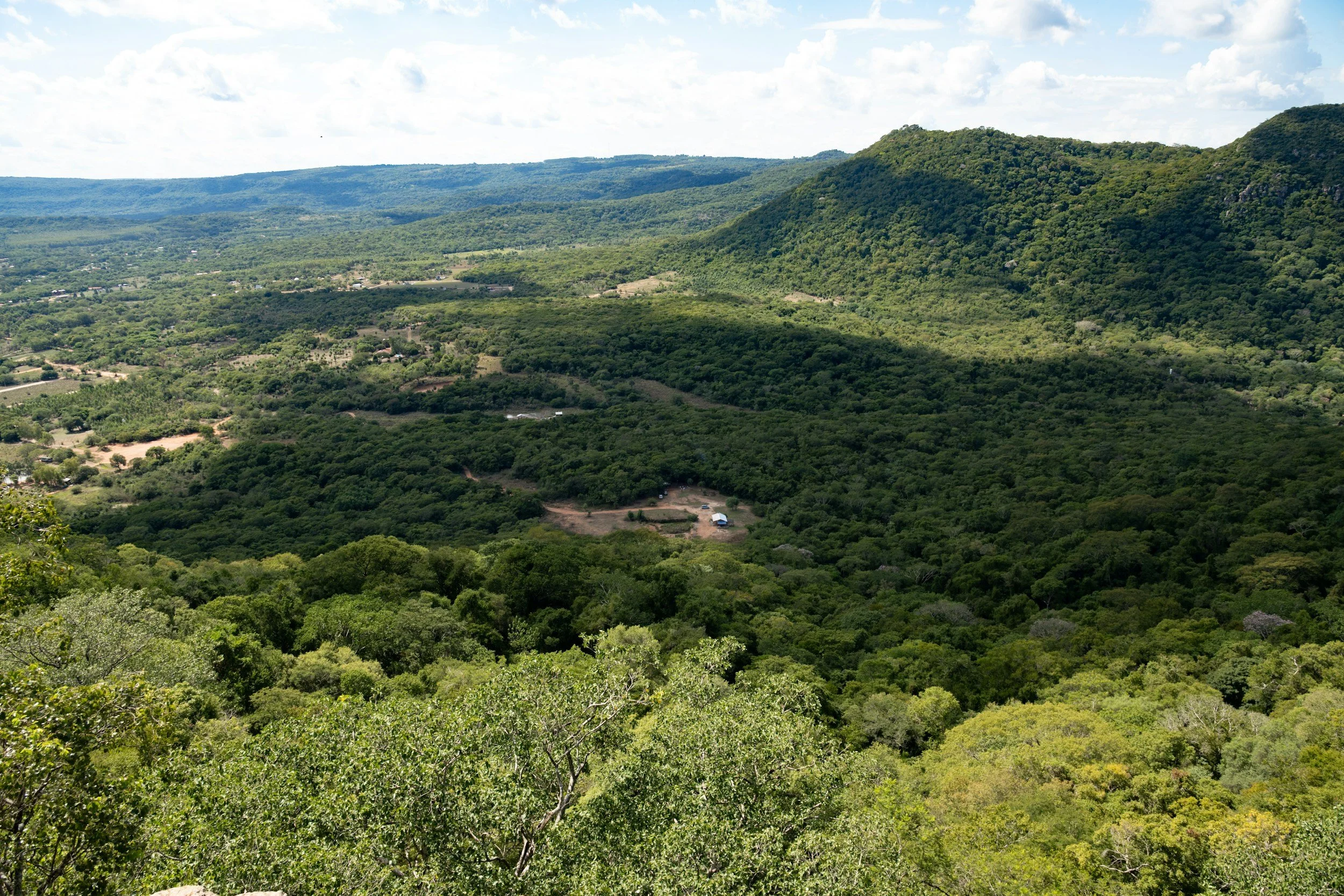 A panoramic view of lush green mountains and valleys with scattered buildings, under a partly cloudy sky.