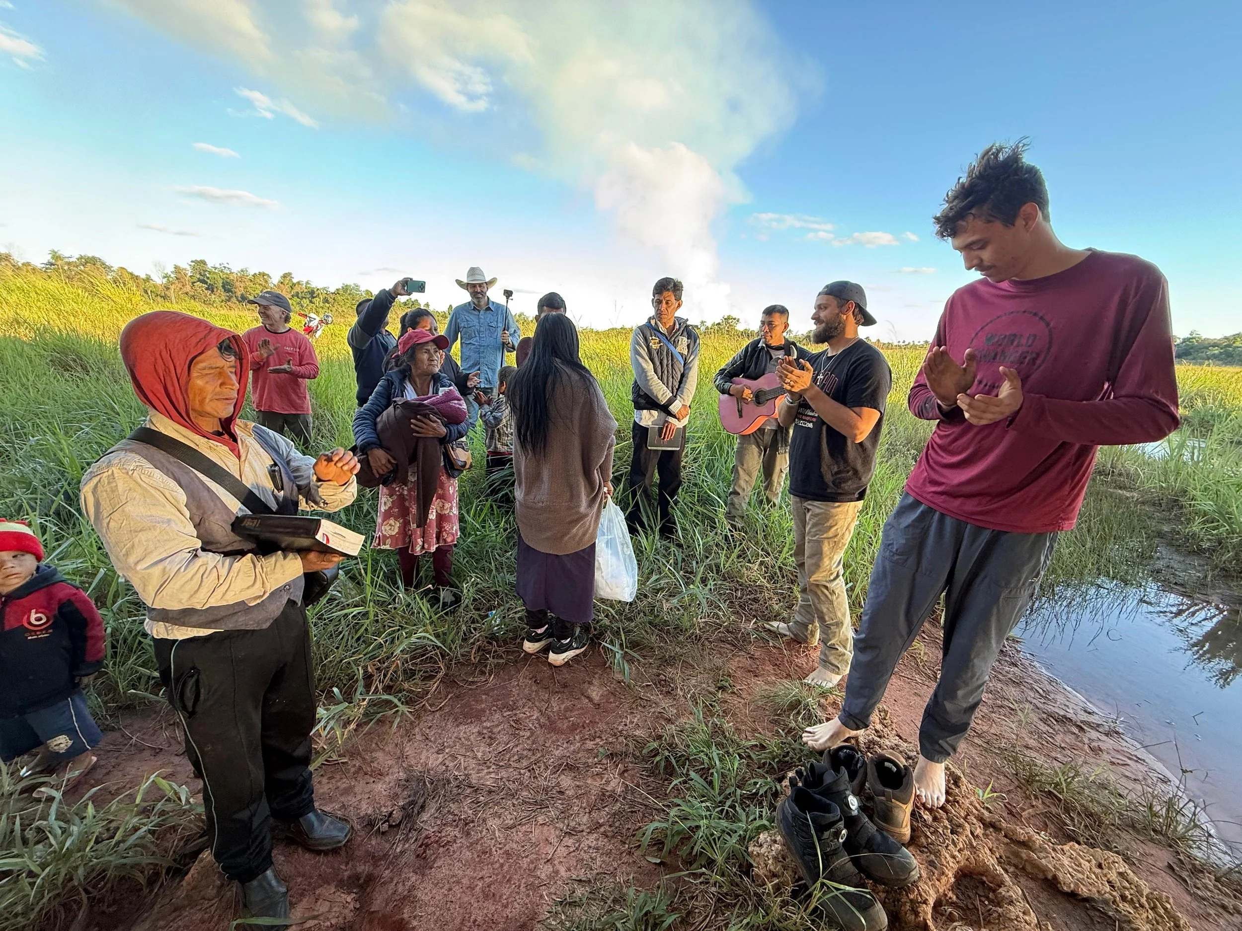 Group of people gathered near a body of water in a grassy area, some standing and some sitting, with a young man in the foreground praying. The background features a blue sky with scattered clouds and some trees.