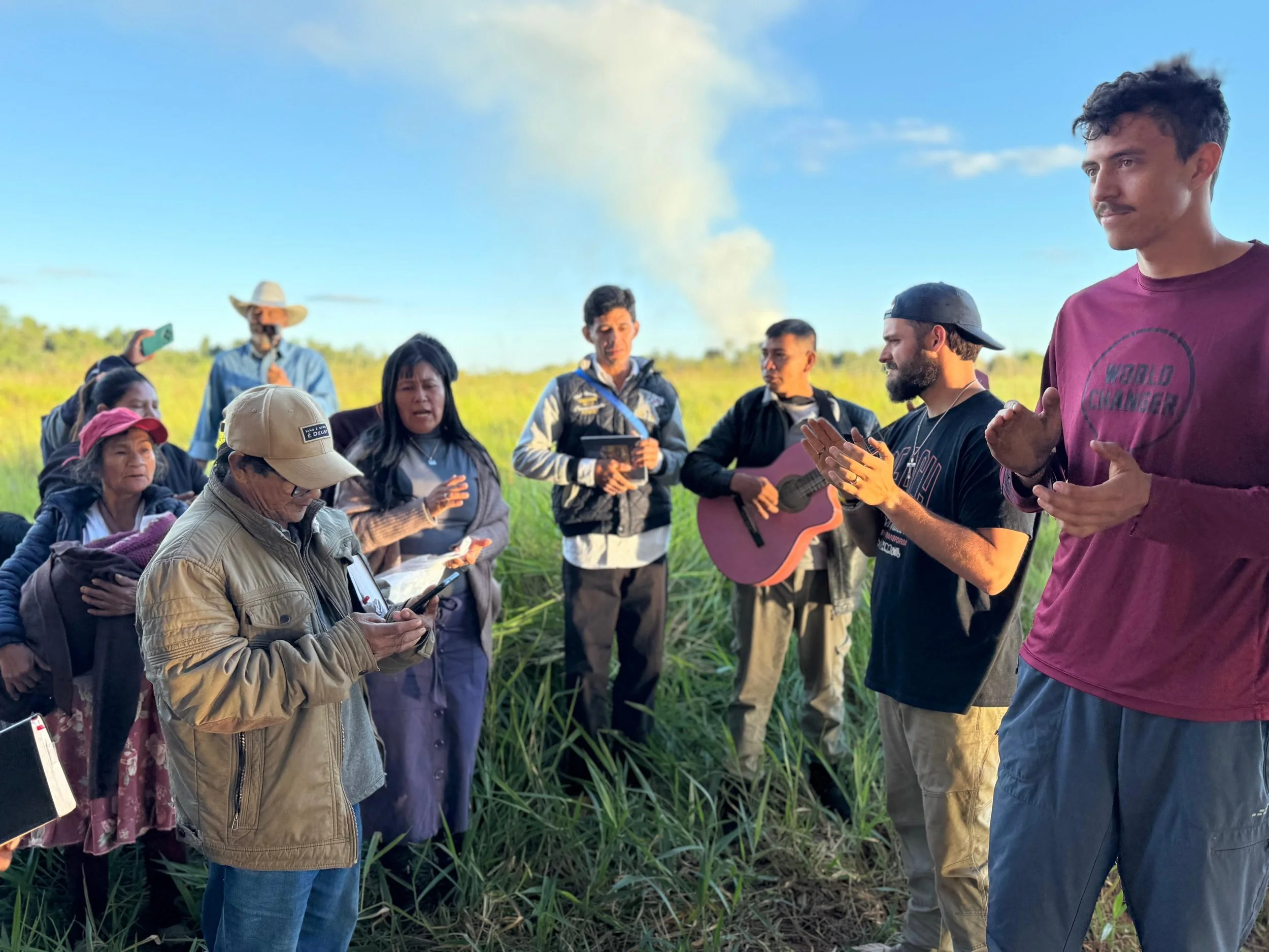 Group of people gathered outdoors in a grassy field, some holding notes and musical instruments, while others participate in a prayer or song, with a clear blue sky and smoke in the background.