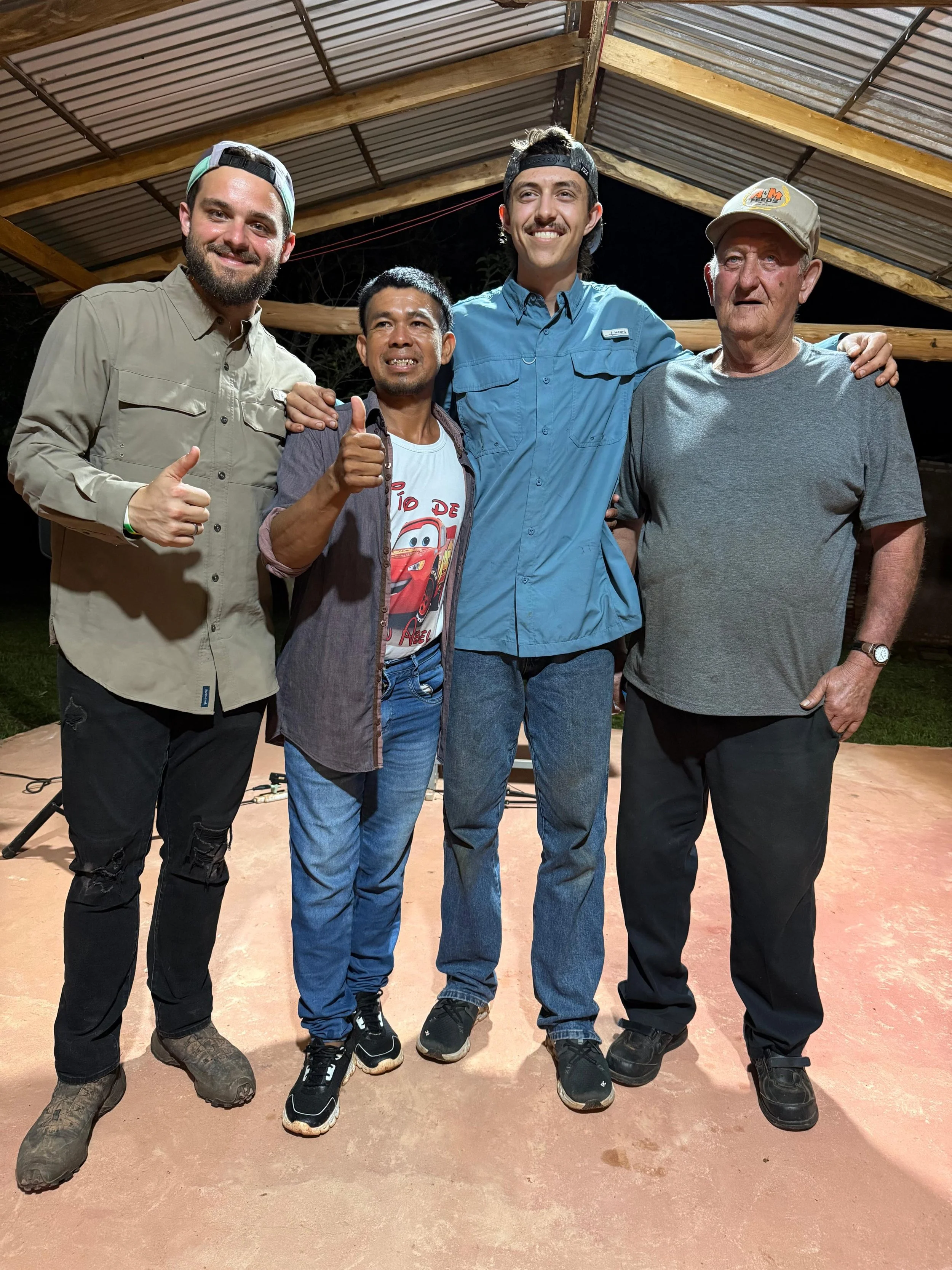 Four men standing together under a metal roof at night. They are smiling and posing for the camera. The man on the far left is giving a thumbs up and wearing a beige shirt and a backward cap. The second man is giving a thumbs-up and wearing a graphic t-shirt, a gray jacket, and jeans. The third man is smiling widely, wearing a light blue shirt and a backwards cap. The man on the far right is older, wearing a gray t-shirt, a beige cap, and a watch.