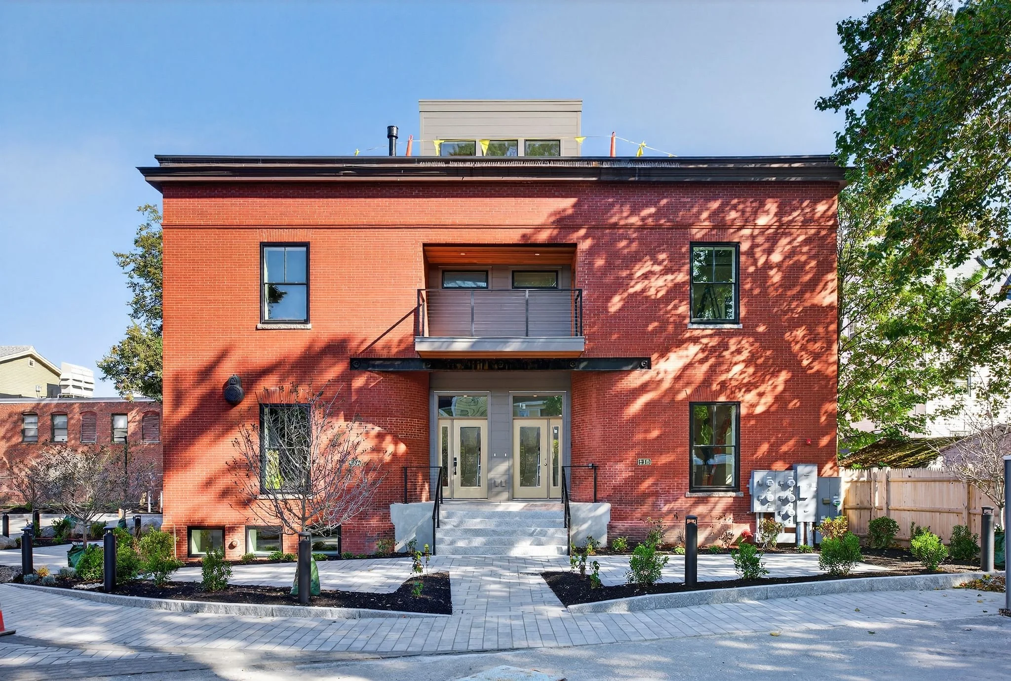 A red brick multi-story residential building with a small balcony on the second floor, large windows, and an entrance with steps and a glass door. There is a paved front yard with young plants, black posts, and a garden bed, surrounded by trees and a