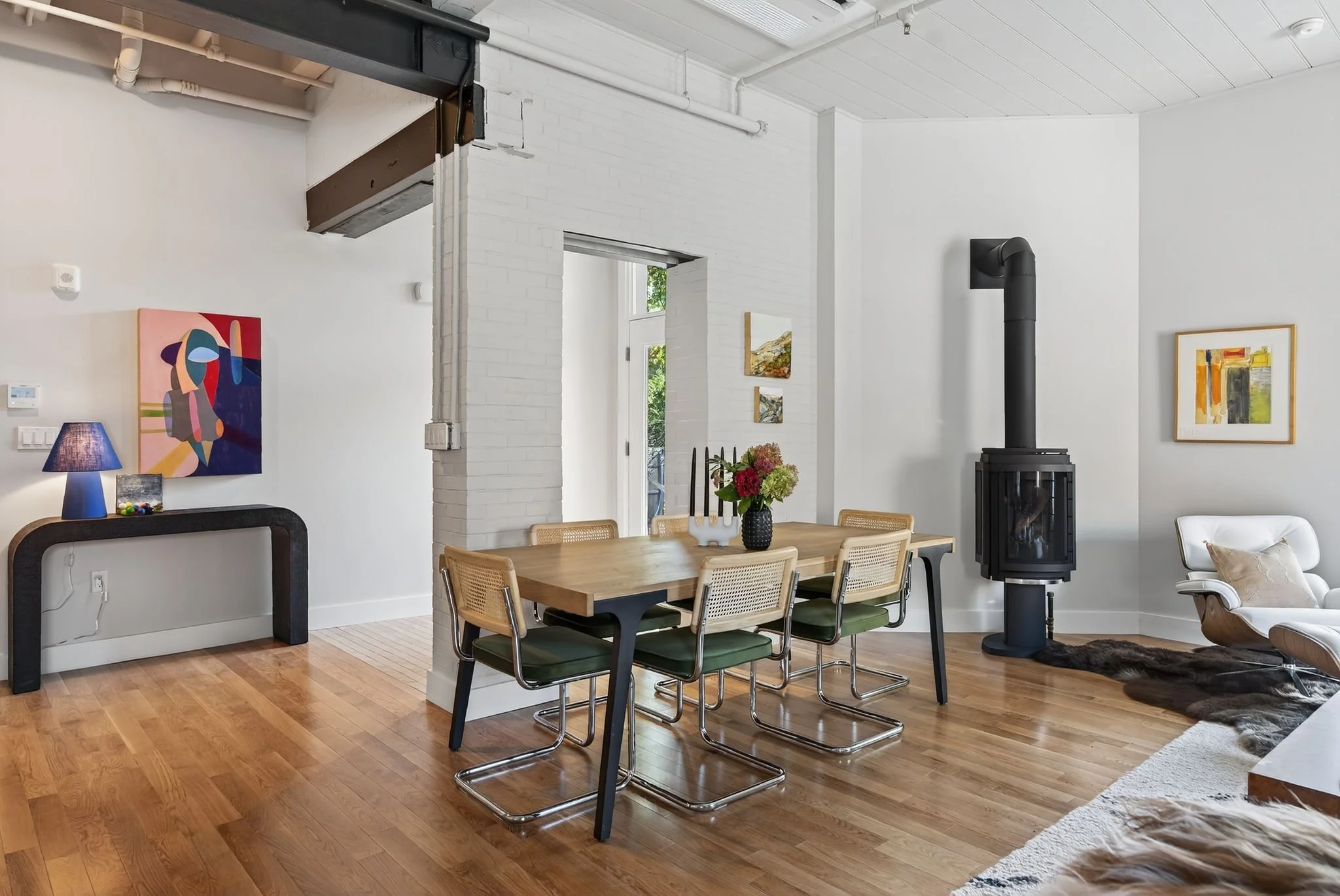 Interior of a bright, modern living and dining area with white walls, wooden flooring, an art piece on the wall, a dining table with chairs, a black fireplace, and a smaller seating area with a white armchair and a rug.