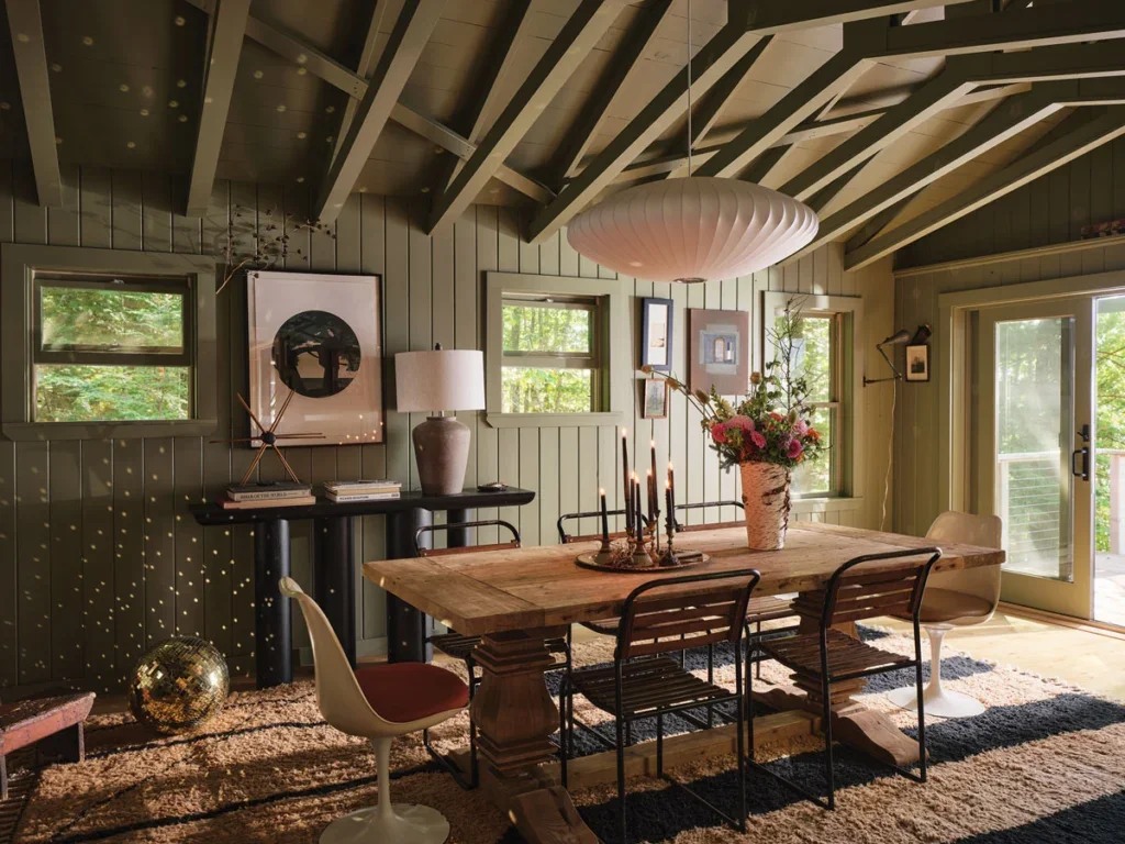 A dining room with a wooden table, floral centerpiece, black candles, and six chairs in front of windows and a door with a balcony. The room has wood paneling, a sloped ceiling with exposed beams, and a large paper lantern light fixture.
