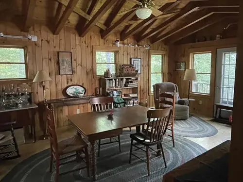 A cozy wooden dining room with a large table, chairs, armchair, bookshelf, and windows showing greenery outside.