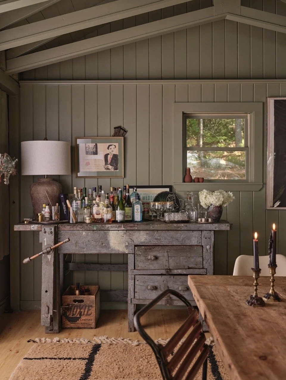 A rustic interior room with a wooden bar cart filled with liquor bottles, a framed picture, a white lamp, and a small plant on top. There is a small window with outside greenery, a wooden table with lit candles, and a beige chair. A beige rug with black lines lies on the wooden floor.