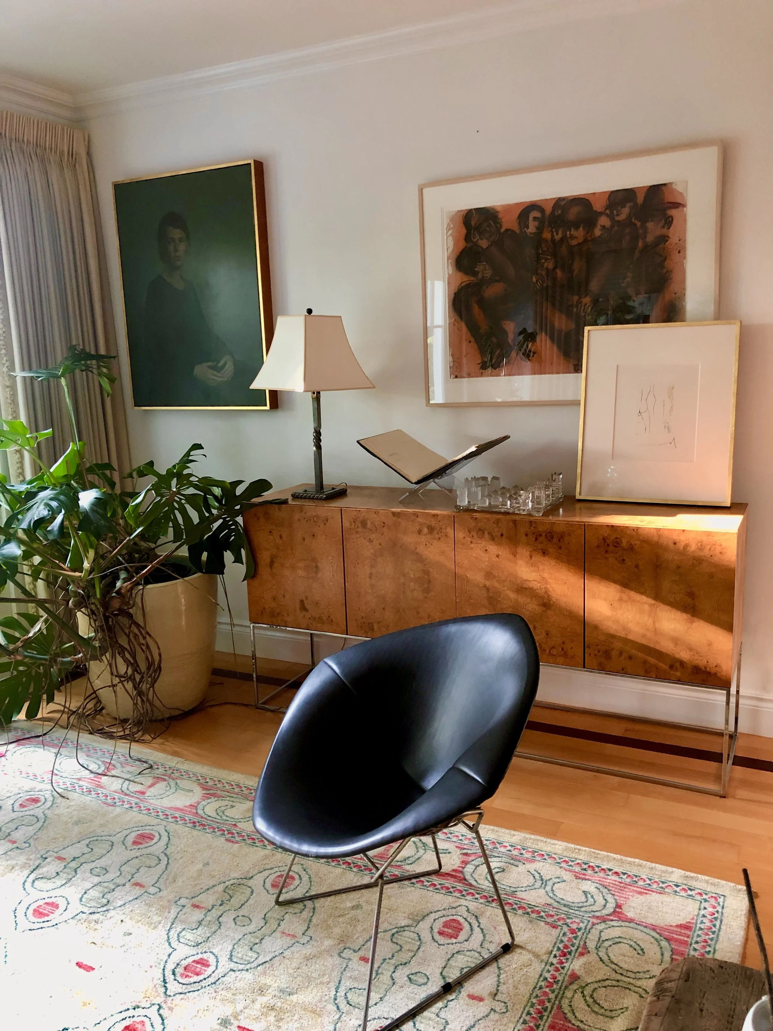 Living room with a large potted plant, a black modern chair, a wooden cabinet with a lamp, framed artwork, and a rug, against beige walls with artwork and curtains.