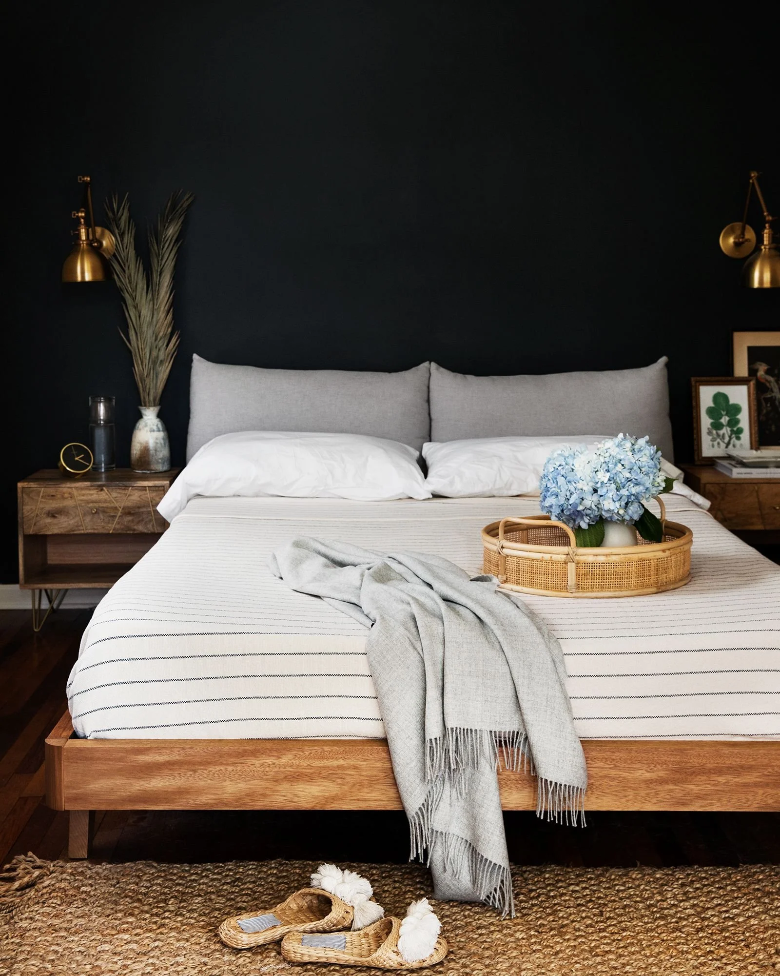 A neatly made bed with a gray headboard and white linens. A light gray throw blanket drapes over the foot of the bed. On the bed, there is a round wicker tray with a blue hydrangea flower arrangement. Fluffy white slippers are on the woven rug at the foot of the bed.
