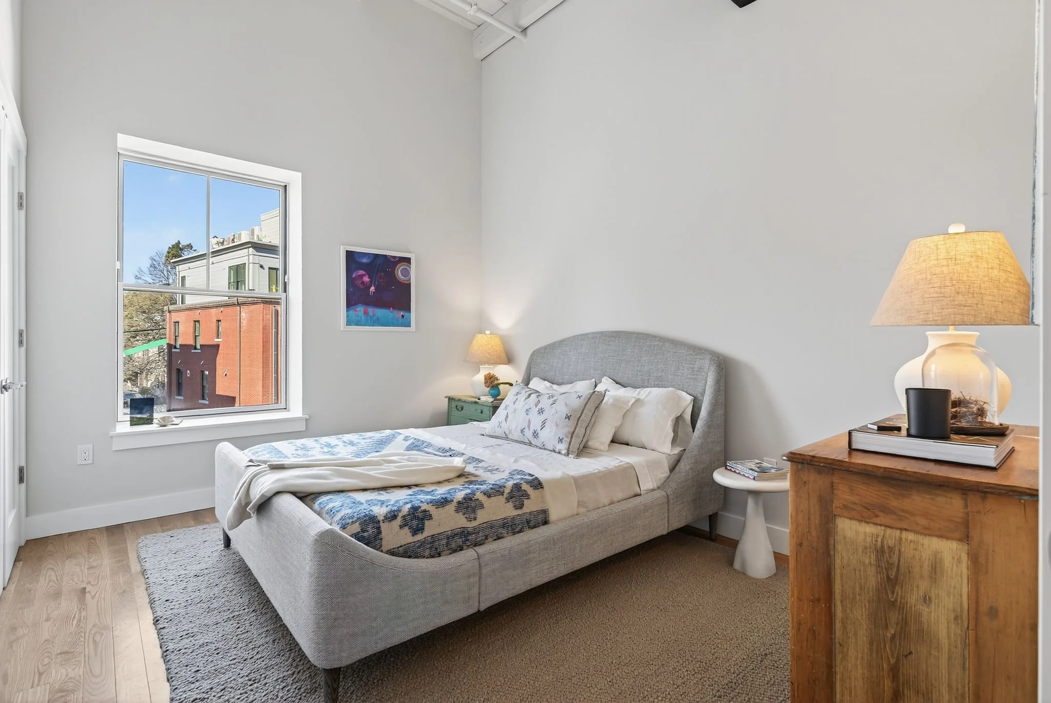 A bedroom with a gray upholstered bed, white linens, and patterned pillows. A nightstand with a lamp, books, and a black speaker. A window showing nearby buildings and trees. Abstract art on the wall.