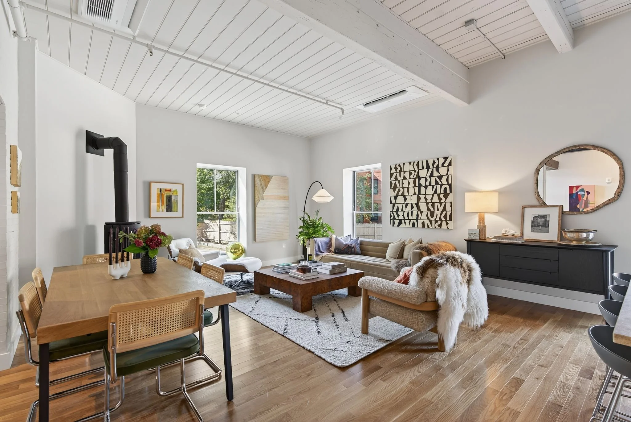 Living room with wooden flooring, white walls, and a white ceiling with exposed beams. Large windows let in natural light. There is a beige sectional sofa with throw pillows, a wooden coffee table, and wall art. A black sideboard has framed photos, a