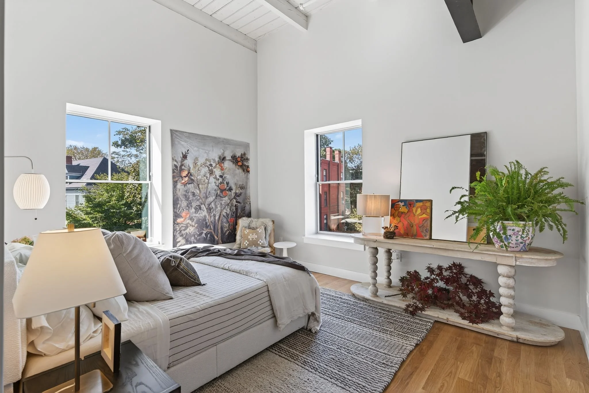 A cozy living room with white walls and high ceiling. There are two windows letting in natural light, decorated with artwork and houseplants. A sofa with cushions and throws is placed near a side table with a lamp, and a rustic white console table ho