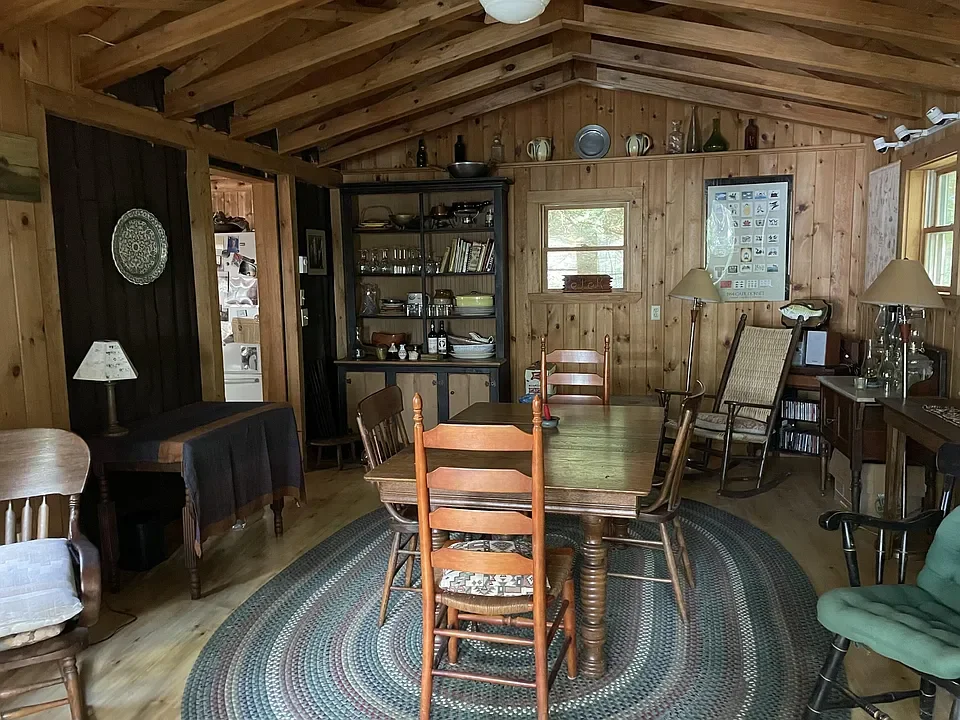 Wood-paneled dining room with a central wooden table surrounded by mixed chairs, a braided rug, shelves with dishes and bottles, and various lamps and decorations.