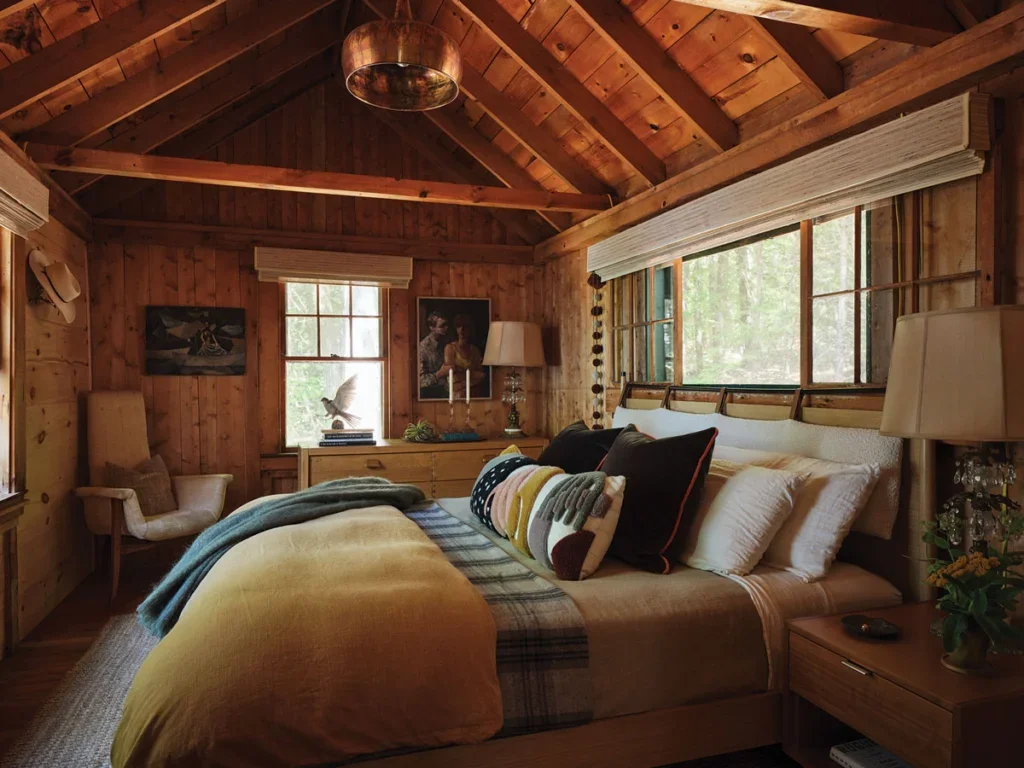 A cozy bedroom with wooden walls and ceiling, a large window with shades, a bed with multiple pillows, and a side table with a lamp and flower vase.