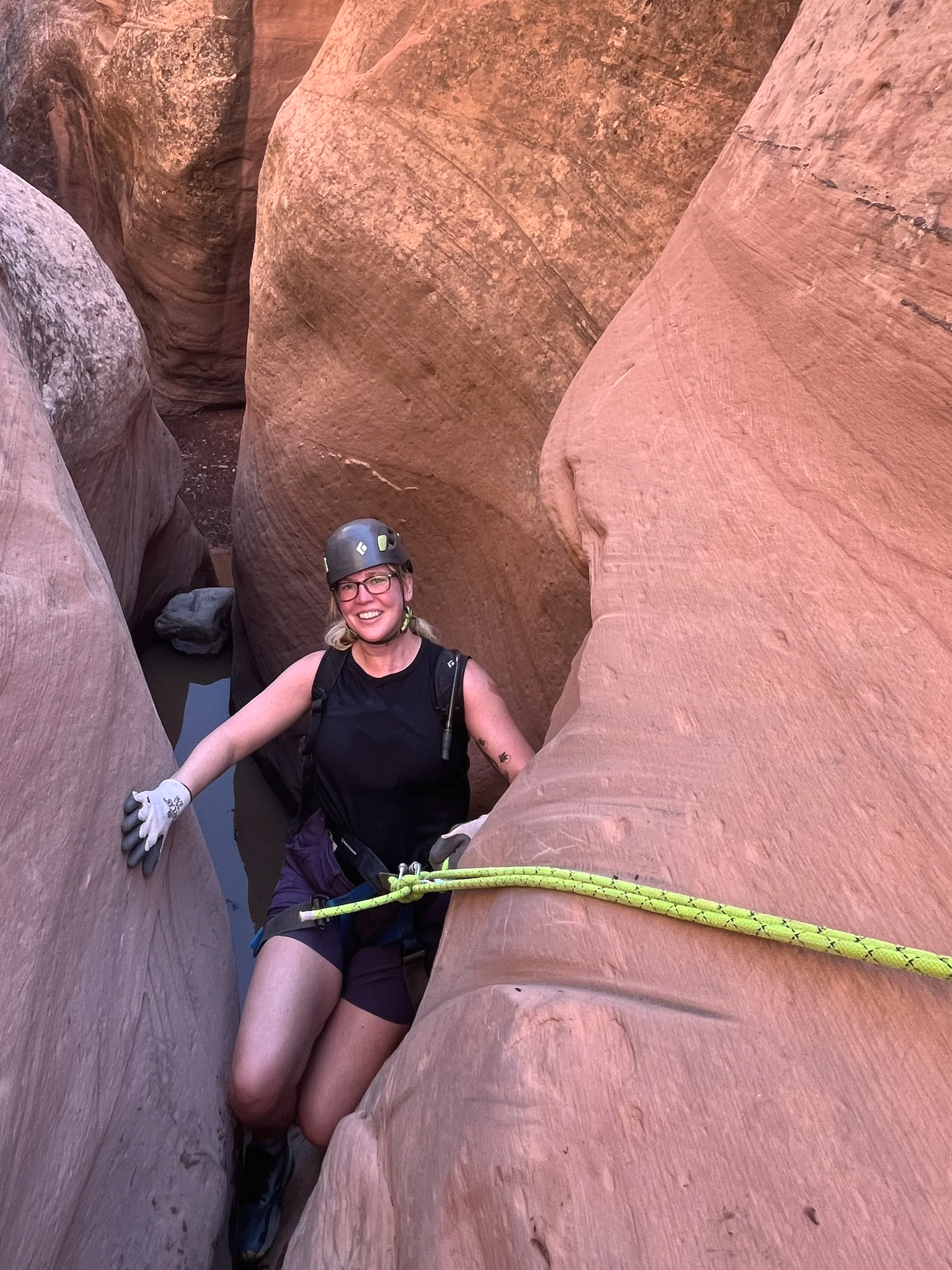 A woman wearing a helmet and gloves rock climbing on large, smooth, pinkish sandstone formations in a canyon.