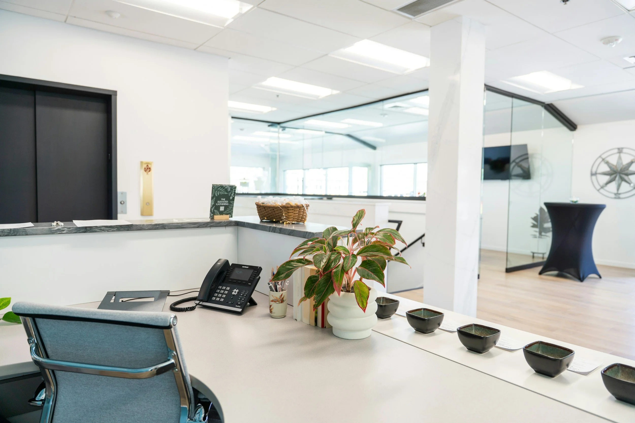 Office reception area with plants on a counter, a computer, a black conference table, black bowls, and glass walls with nautical decor.