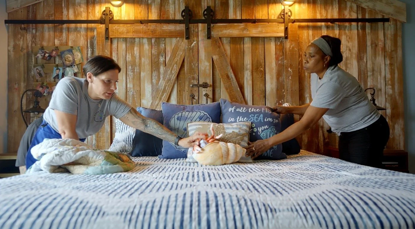 Alexa and cleaning technician are making the bed for residential cleaning service. The bedroom has a wooden barn-style door and rustic decor.