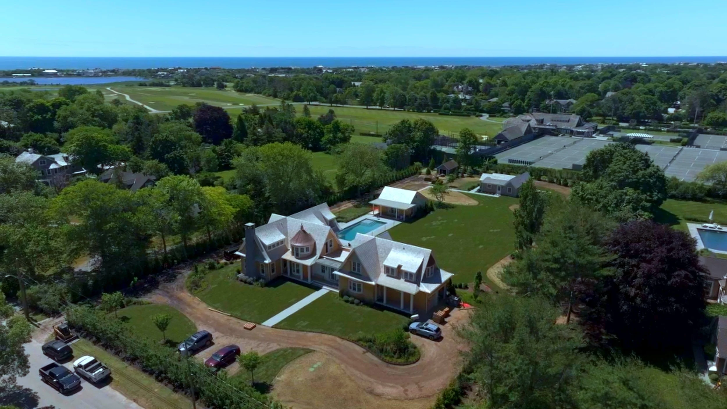 Aerial view of a coastal neighborhood with large houses, trees, a green golf course, and the ocean in the background.