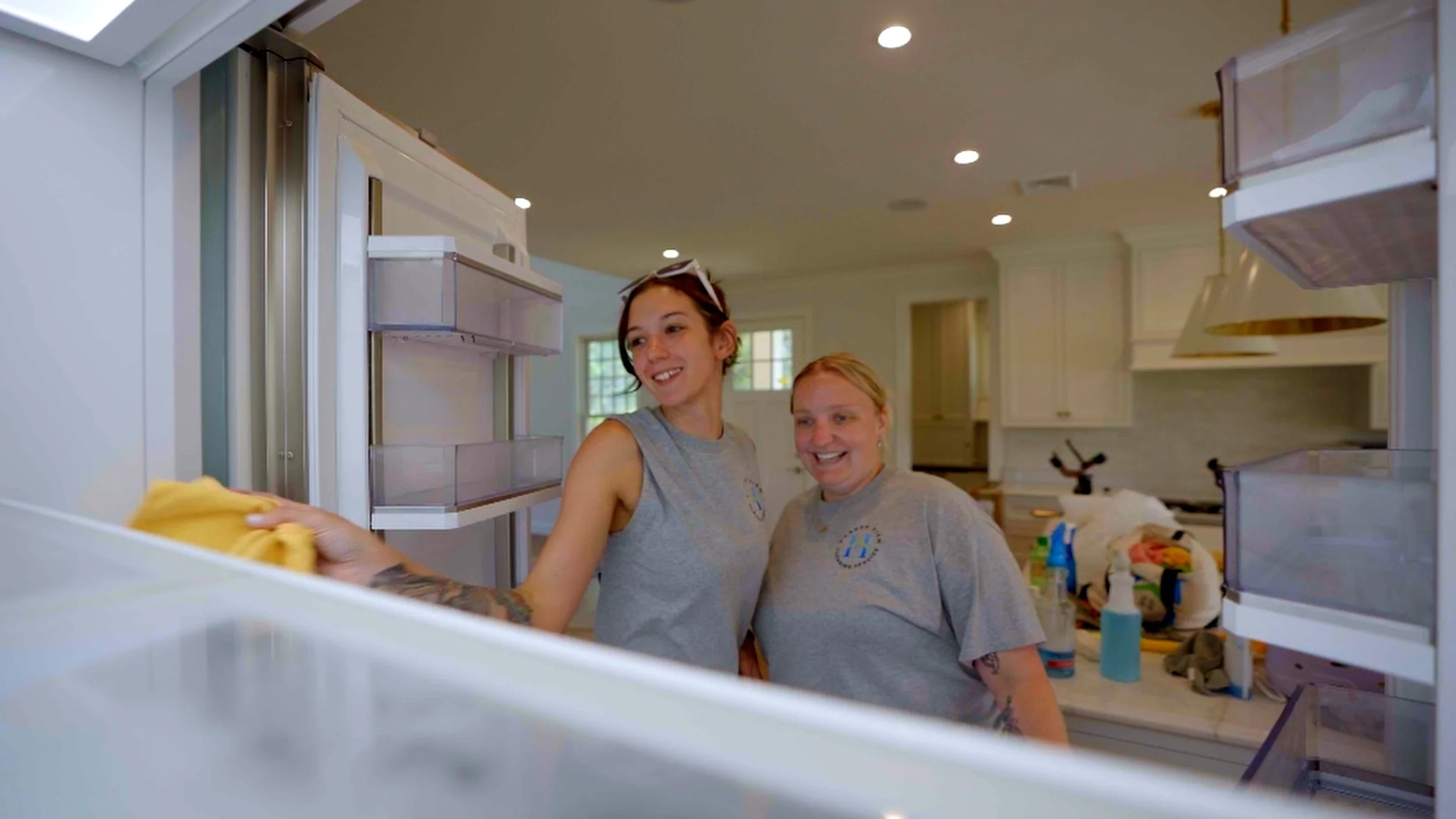 Two women smiling and cleaning the interior of a refrigerator in a bright, modern kitchen.