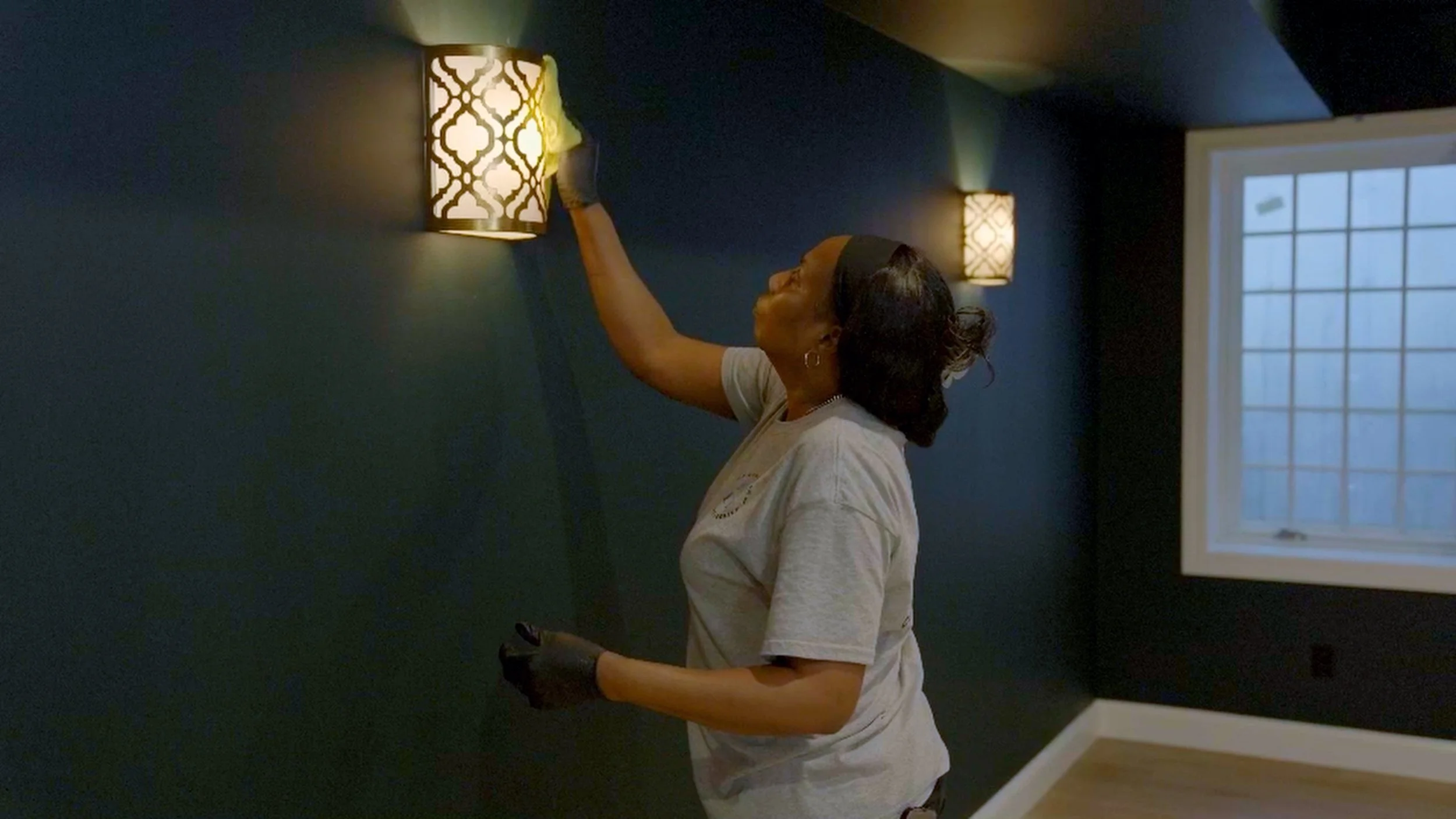 A woman cleaning a decorative wall light fixture in a room with dark walls and a window.