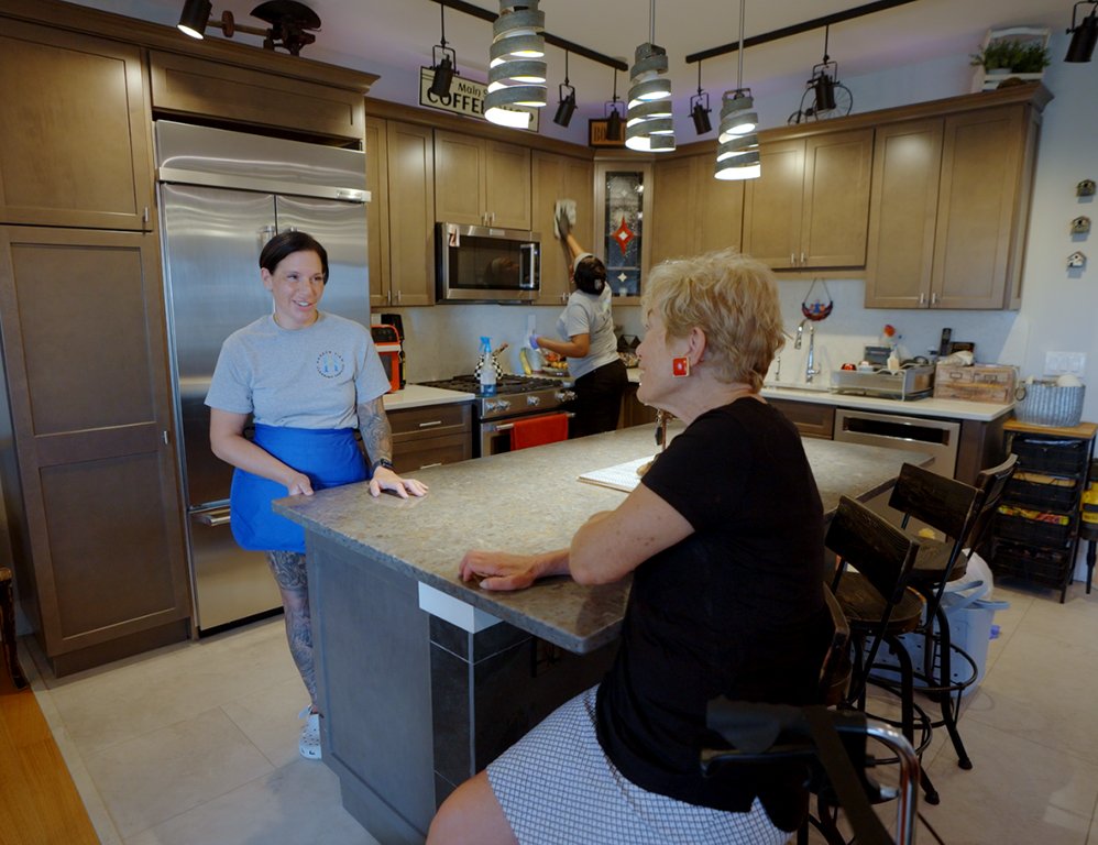 Alexa sits with a client at a kitchen island, speaking with someone behind the counter while another woman cleans in the background of a modern kitchen.