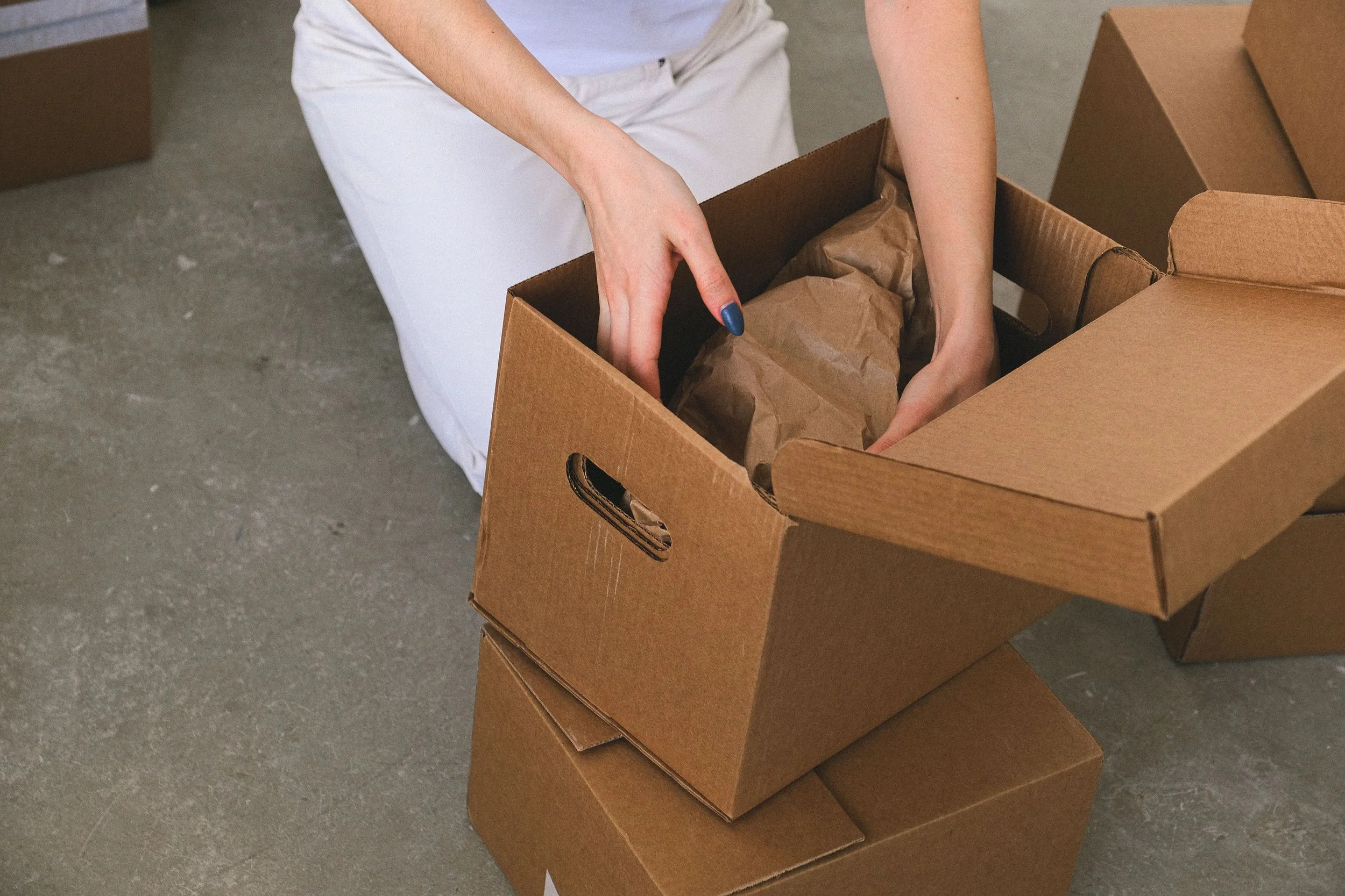 Move in / Move out cleaning services with a person packing brown paper wrapped items into cardboard boxes surrounded by more cardboard boxes on a concrete floor.