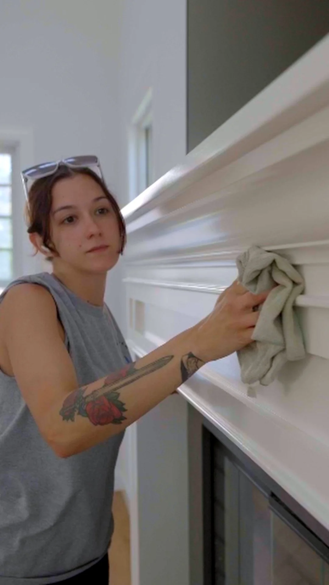 Woman cleaning a white wooden shelf with a cloth, wearing glasses on her head and a sleeveless gray shirt, with tattoo of a flower on her arm, in a well-lit room.