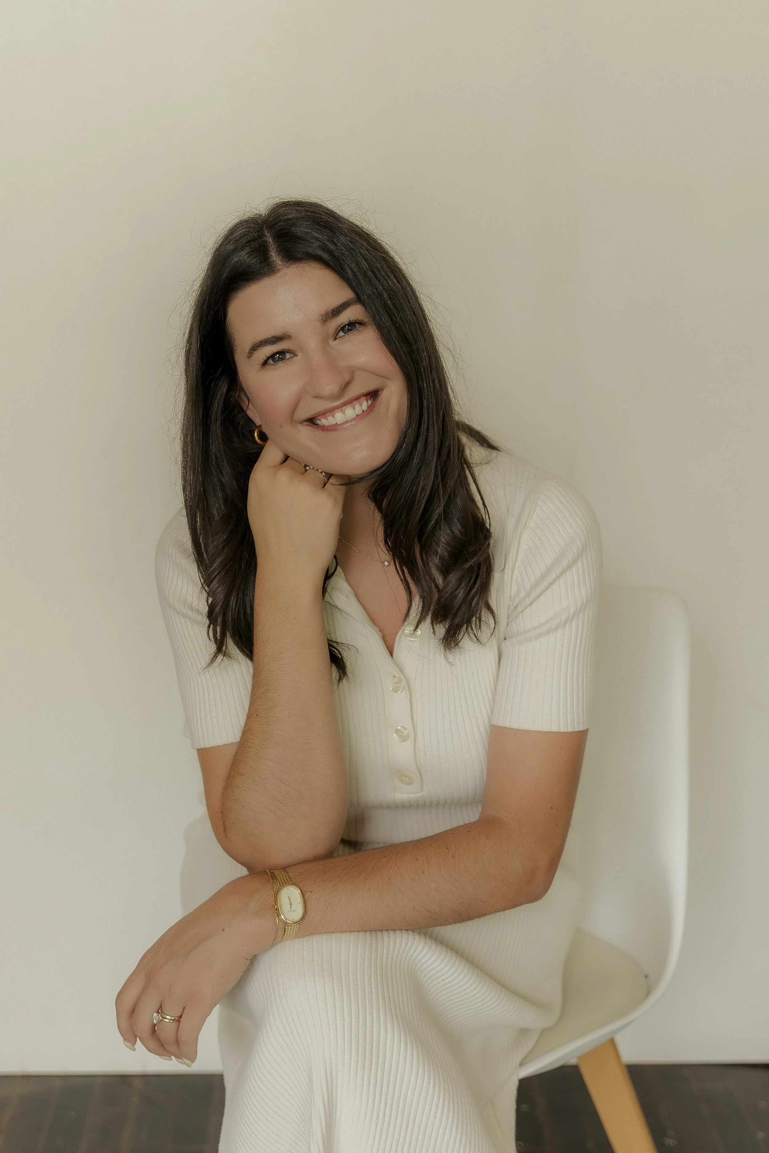 A woman with dark brown hair, smiling, wearing jewelry, a cream-colored short-sleeve button-up top, and sitting on a white chair with a wooden leg, positioned against a plain light-colored wall.