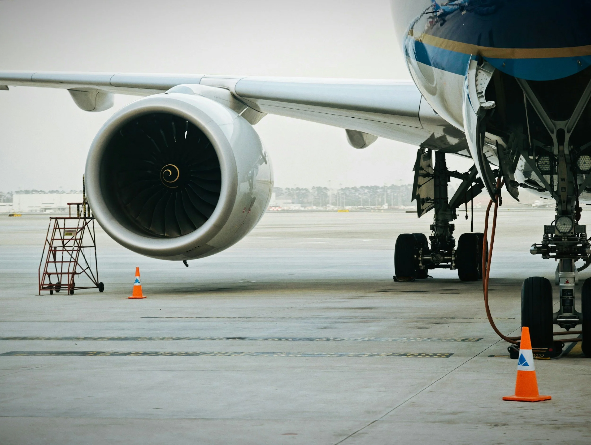 Part of a commercial airplane on the tarmac, showing a large jet engine, landing gear, and runway cones, with airport buildings in the background.