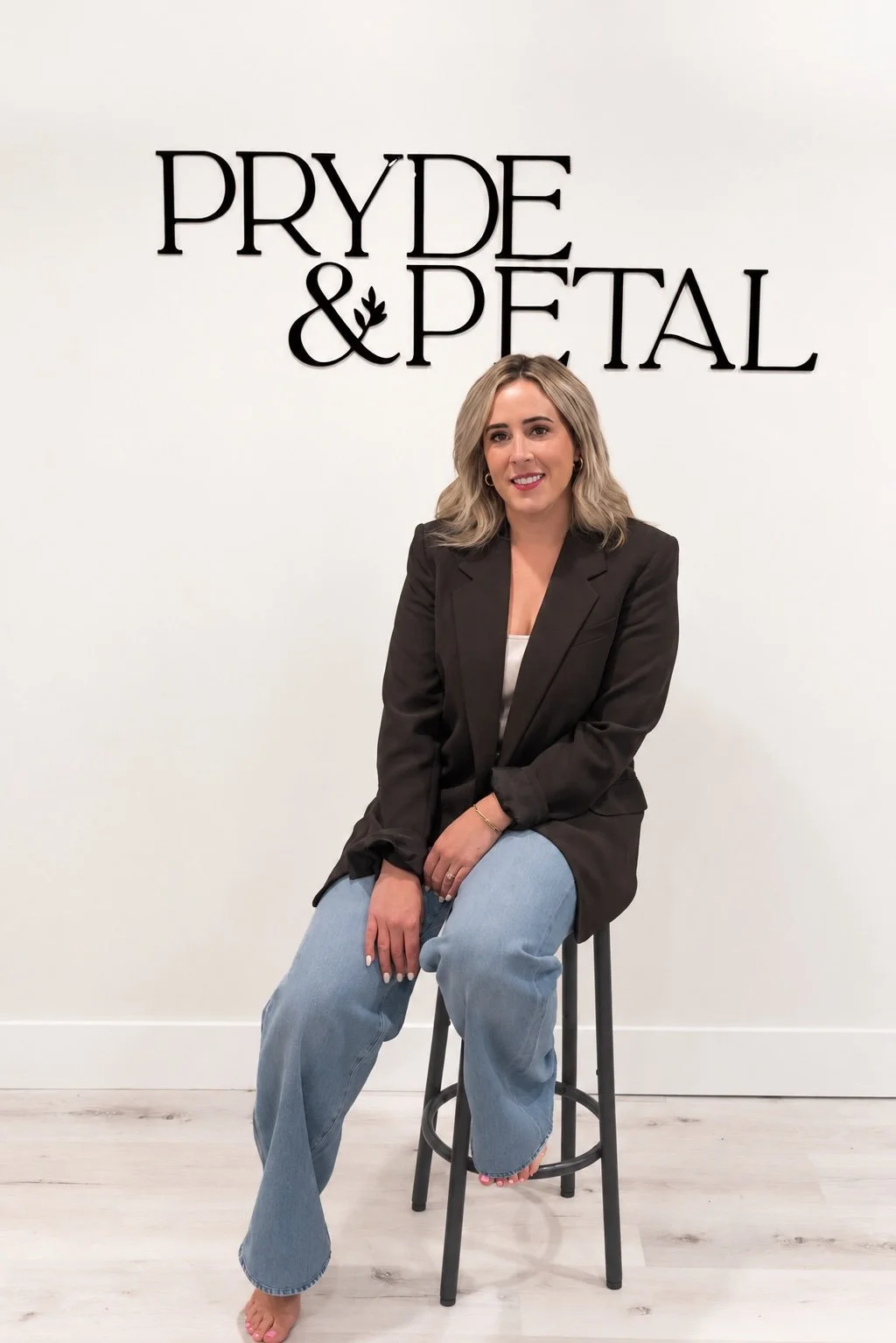 A woman with blonde hair sitting on a black stool in front of a white wall with 'PRYDE & PETAL' written in black lettering.