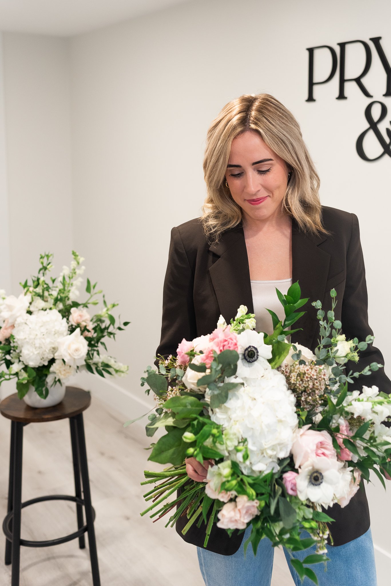 A woman in a brown blazer and blue jeans holding a large bouquet of flowers, with more floral arrangements on a table and a second stand in the background inside floral studio.