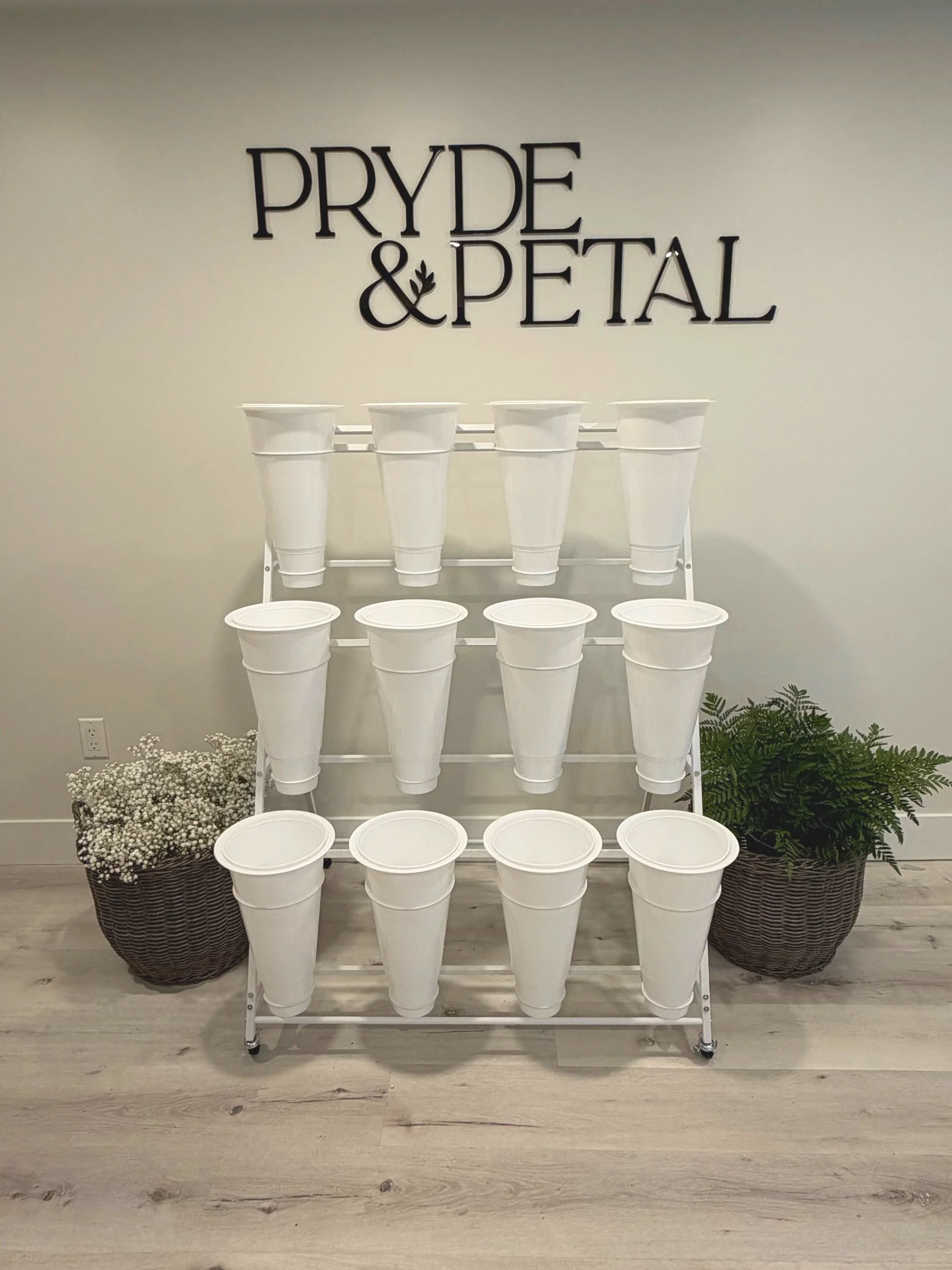 A display of white flower buckets arranged on a white rolling cart with three shelves, positioned between two large woven baskets filled with white baby's breath flowers and green foliage. A wall sign reads 'PRYDE & PETAL' in black lettering.
