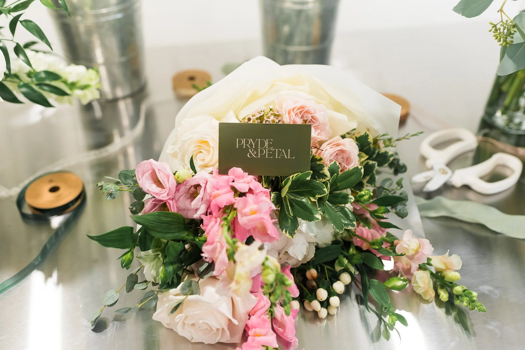 A floral bouquet featuring pink and white roses, accented with greenery and white berries, sitting on a reflective metallic surface with blurred green foliage in the background.