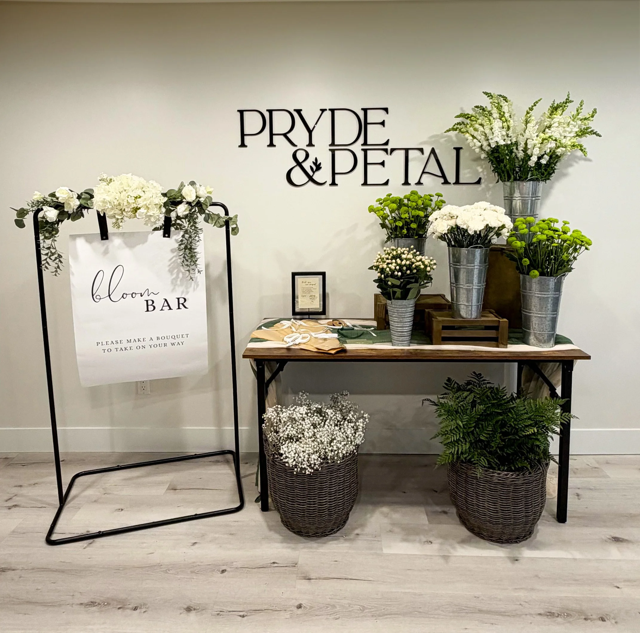 Display of white flowers in vases, a sign for a wedding flower bar, and a decorative board with the words 'bloom BAR' on a white background, arranged on a wooden table.