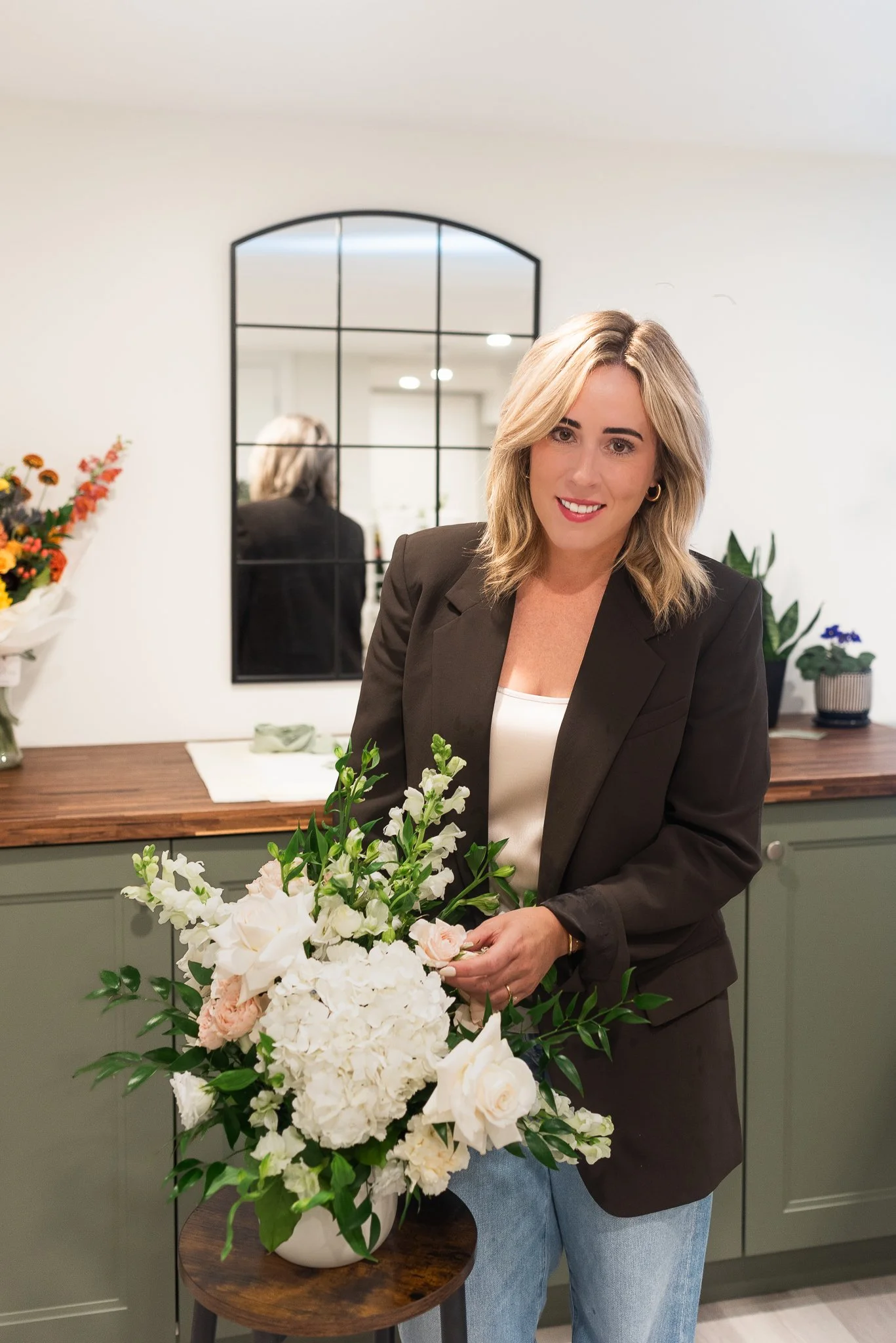 A woman with blonde hair, wearing a brown blazer, stands in a room with a large mirror and a green cabinet, holding a bouquet of white and light pink flowers, smiling at the camera.