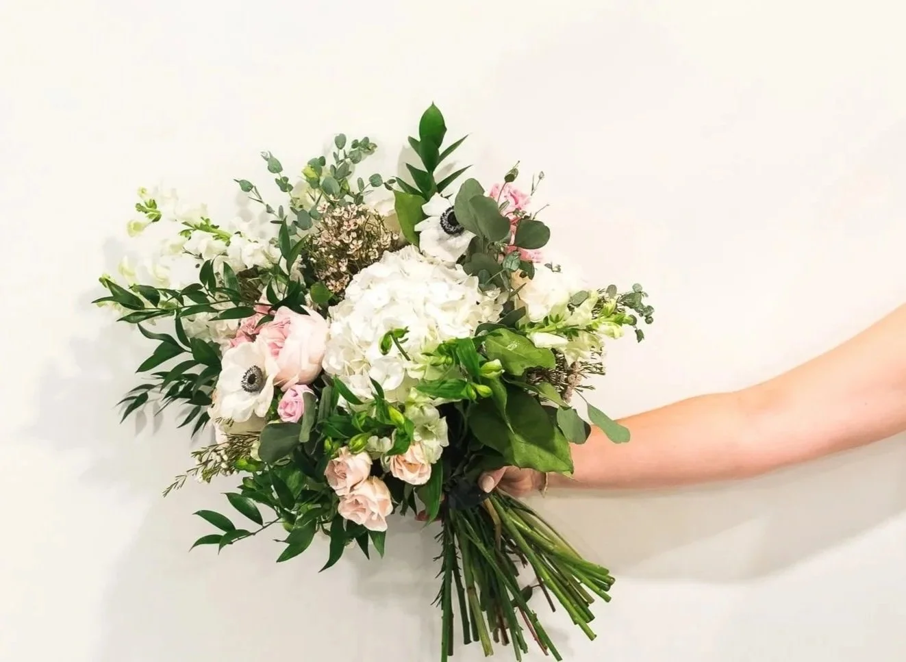 A bouquet of various white, pink, and green flowers and foliage, held by a person's arm stretched out against a plain white wall.