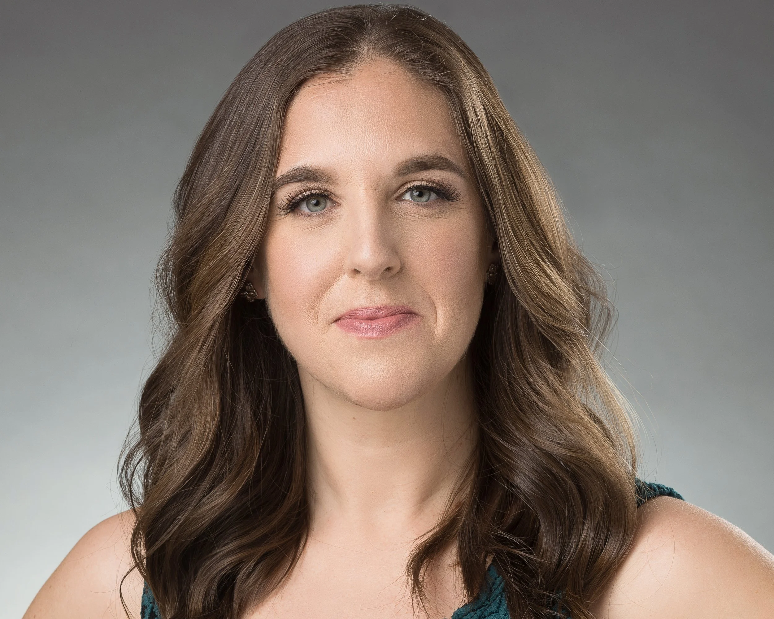 Close-up of a woman with long, wavy brown hair, fair skin, and gray eyes, wearing makeup and a black and blue top, against a gray background.
